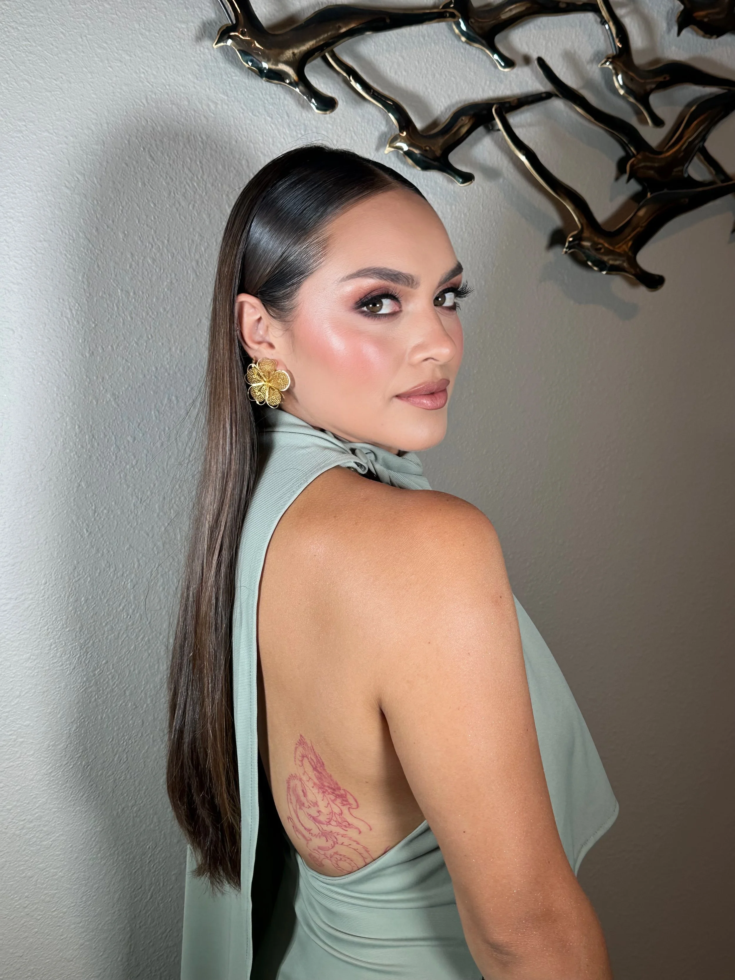 A woman with long dark hair, makeup, and gold flower earrings, wearing a light gray sleeveless top, shows a pink tattoo of a wolf on her upper side.