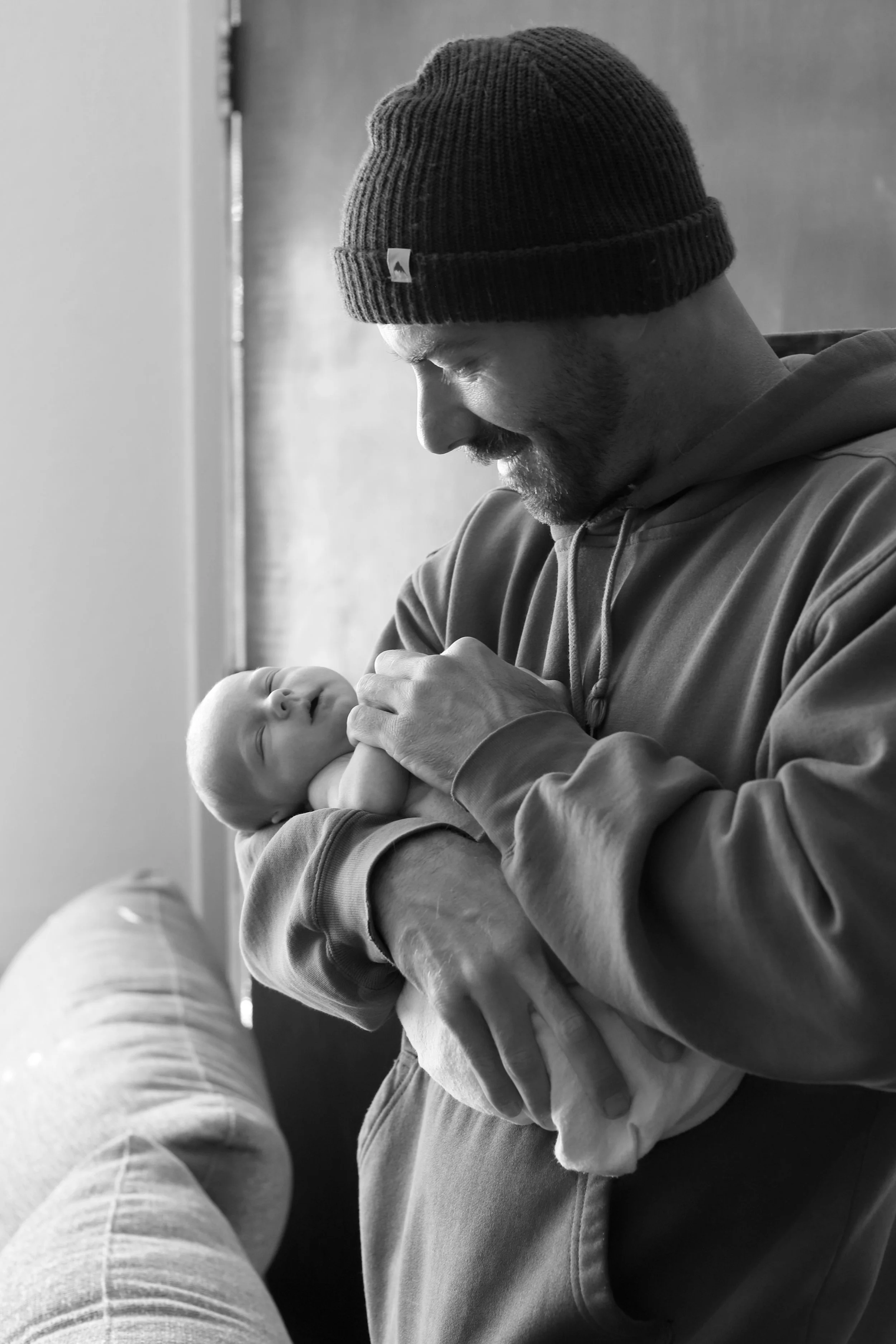 A new father wearing a beanie and hoodie holding his sleeping baby close to his chest, smiling softly.