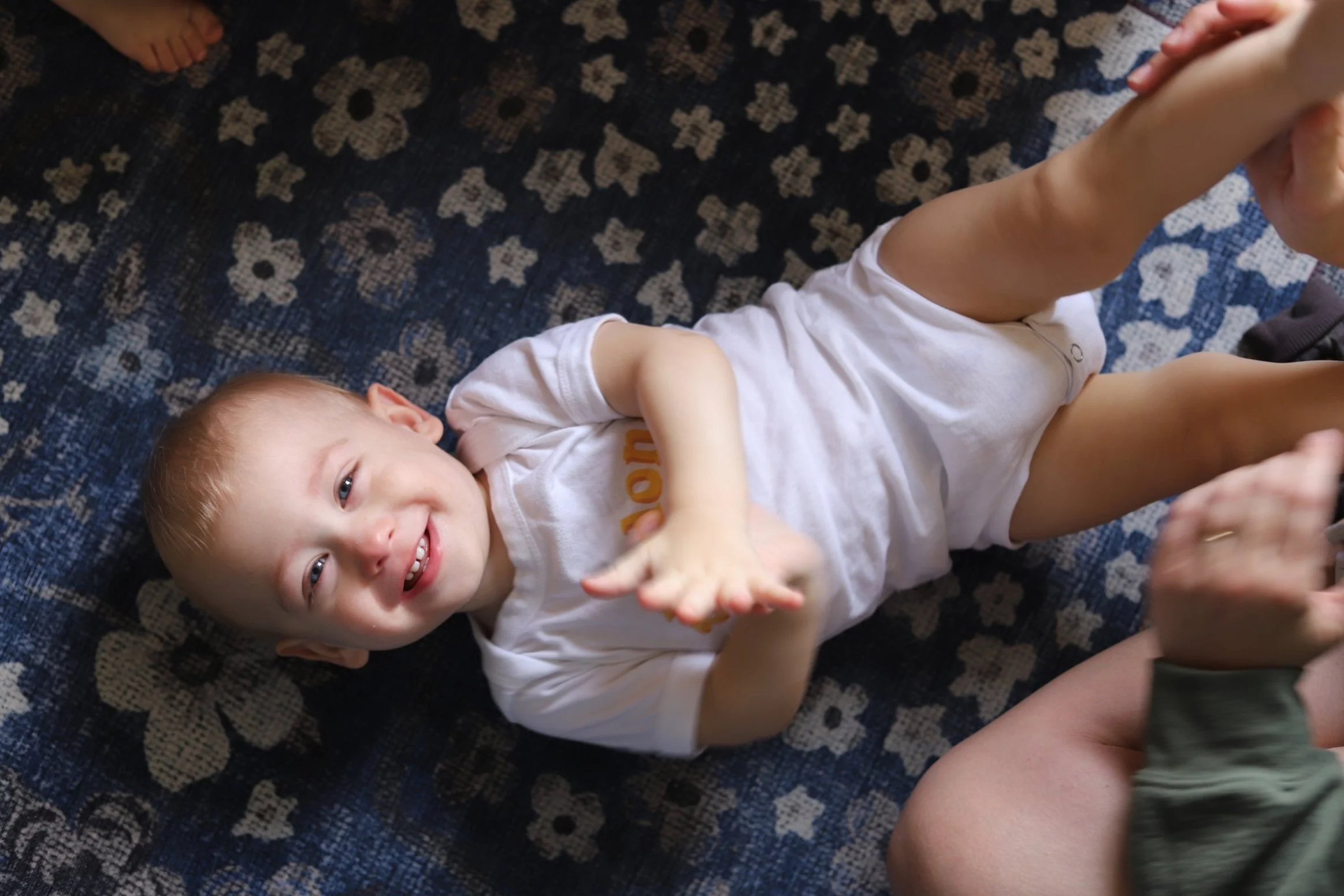 A baby boy lying on a patterned blue carpet, smiling and reaching out with one hand.