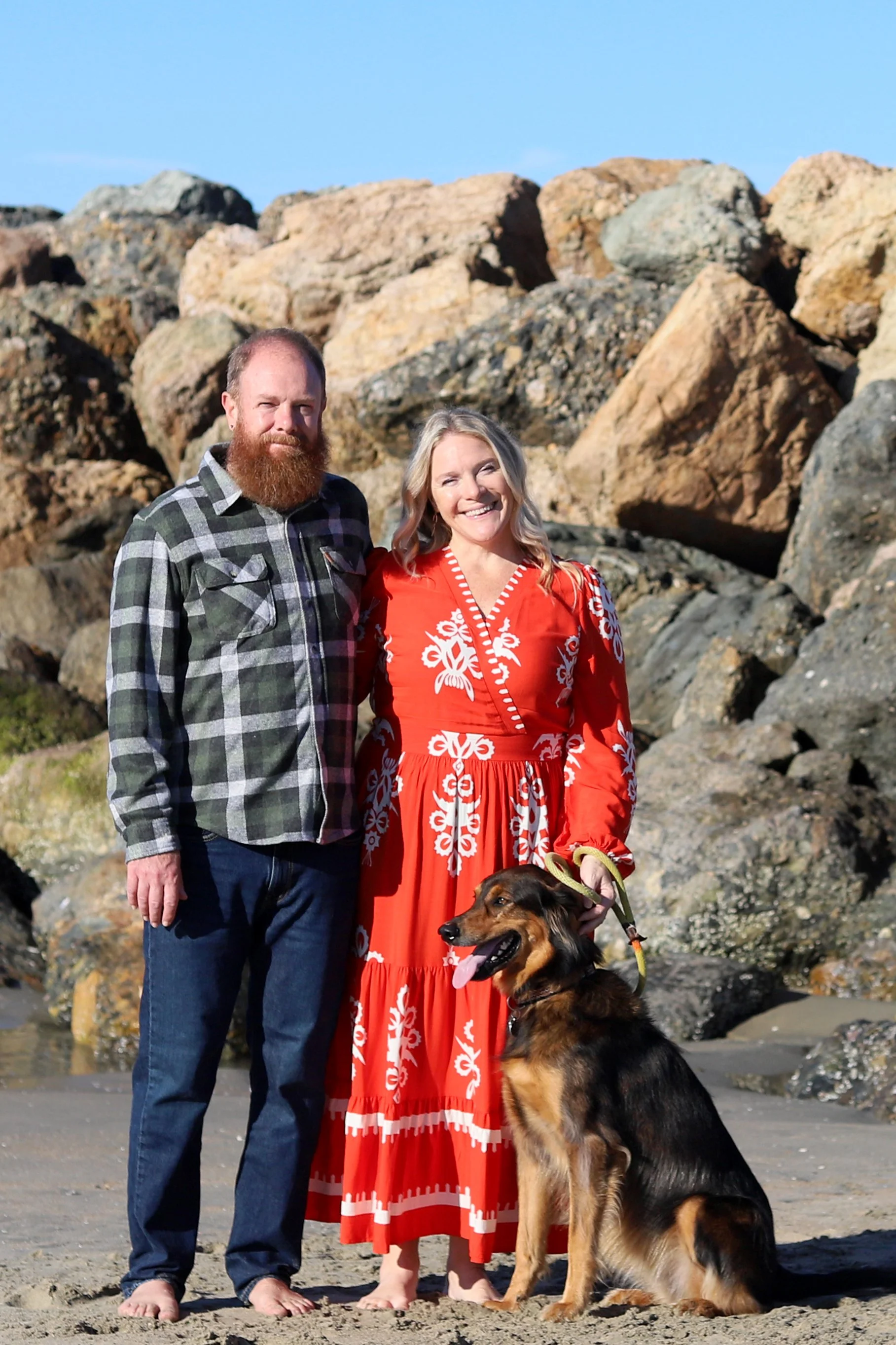 A couple and their dog standing on a beach with large rocks behind them, sunny weather, casual attire.