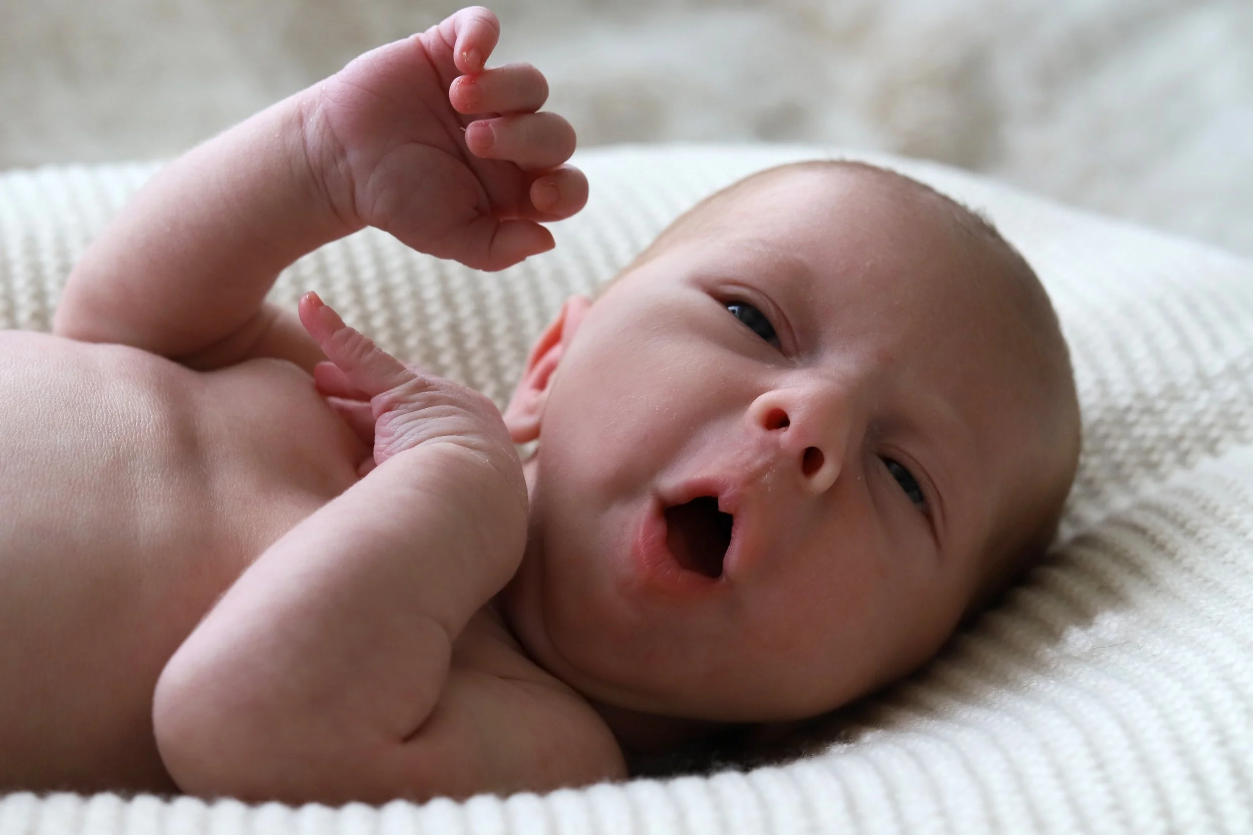 Close-up of a newborn baby lying on a soft surface, with one hand raised and mouth slightly open, eyes partially open.