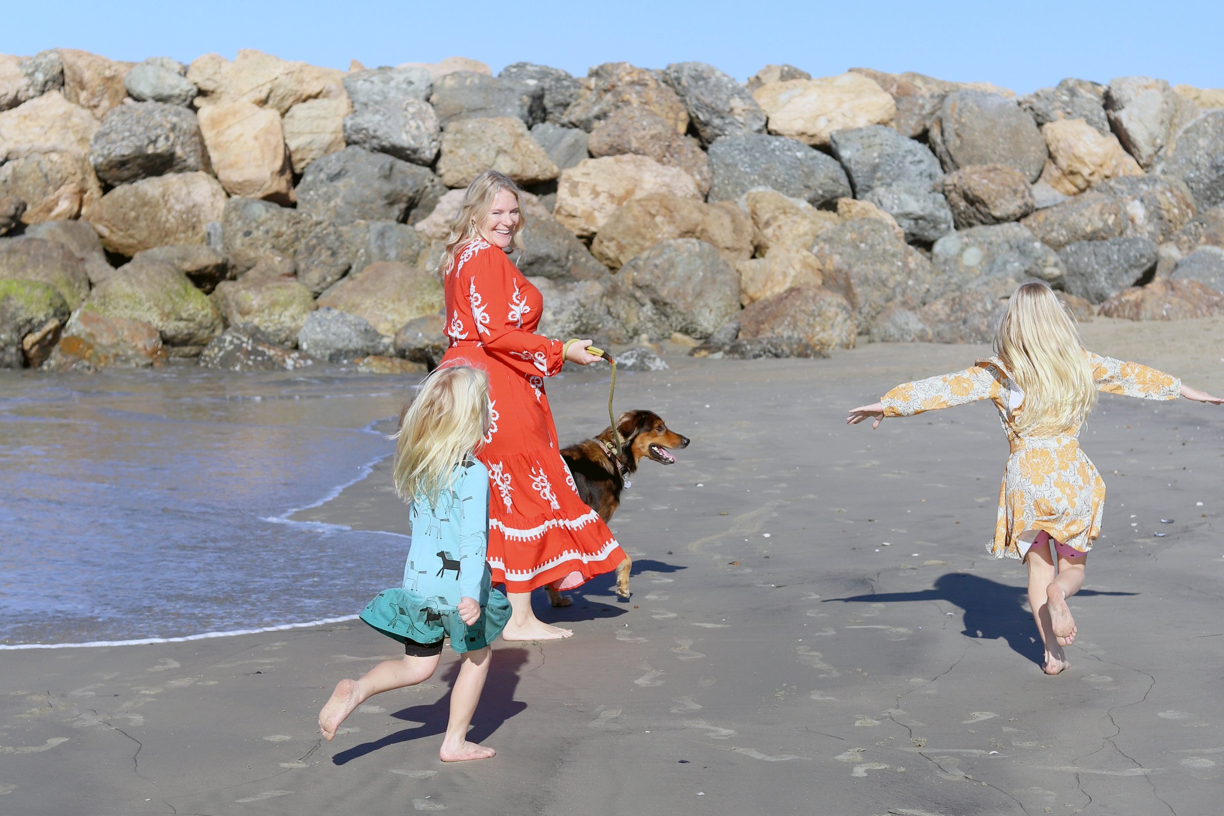 A woman and two young girls are playing on a sandy beach near a rocky shoreline. The woman is holding a leash attached to a dog, and the girls are running with their arms outstretched in a joyful manner. The scene is sunny with clear skies.