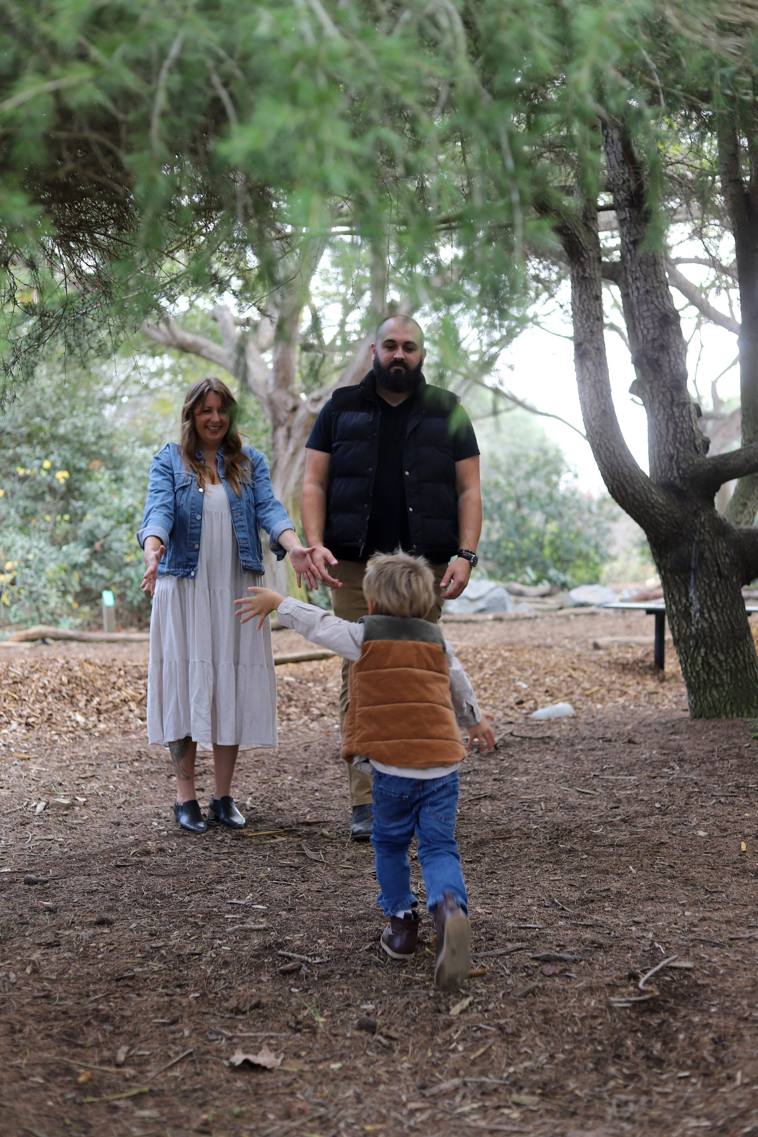 A family of three, a woman and a man with a young boy, is outdoors in a forest clearing. The woman is smiling and reaching out to the boy as he runs toward her, while the man stands nearby observing. The scene is framed by trees and natural foliage.