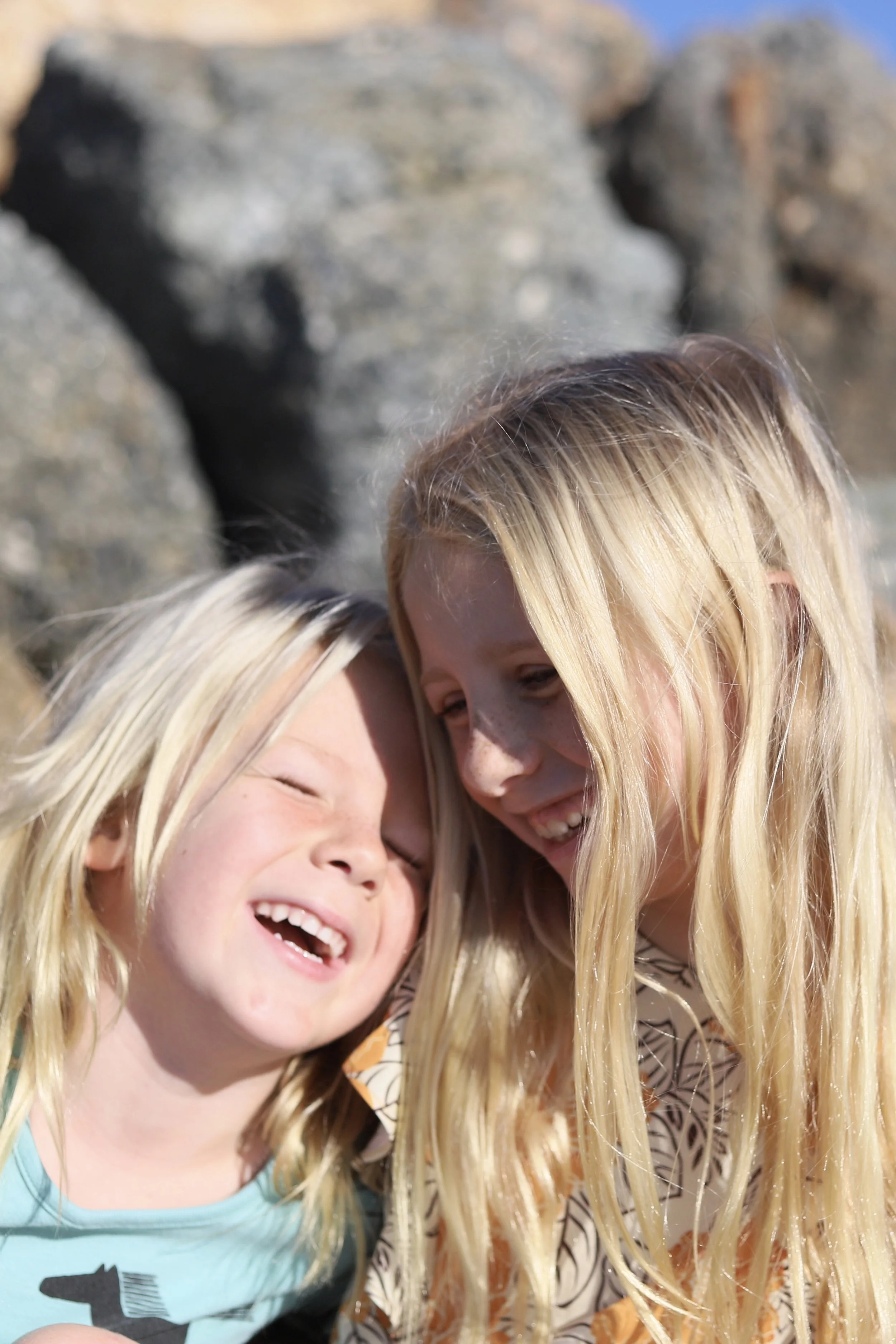 Two young blonde girls smiling and leaning their heads together on a sunny day with a rocky beach in the background.