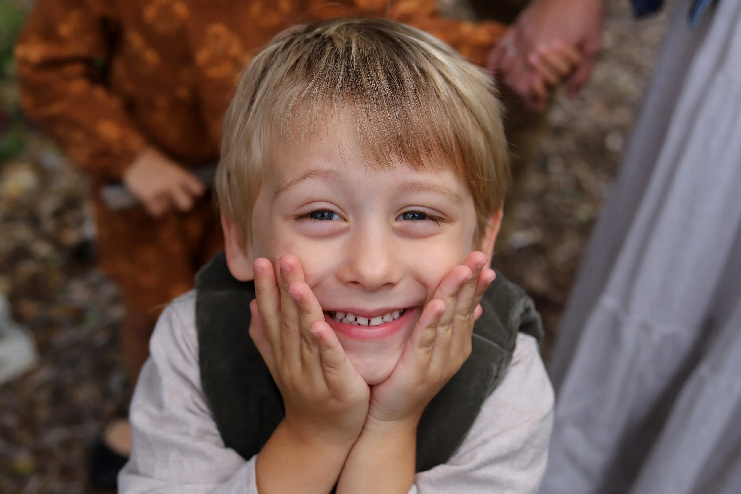 A young boy smiling with hands on his cheeks, wearing a light-colored shirt and vest, outdoors on a dirt ground with fallen leaves.