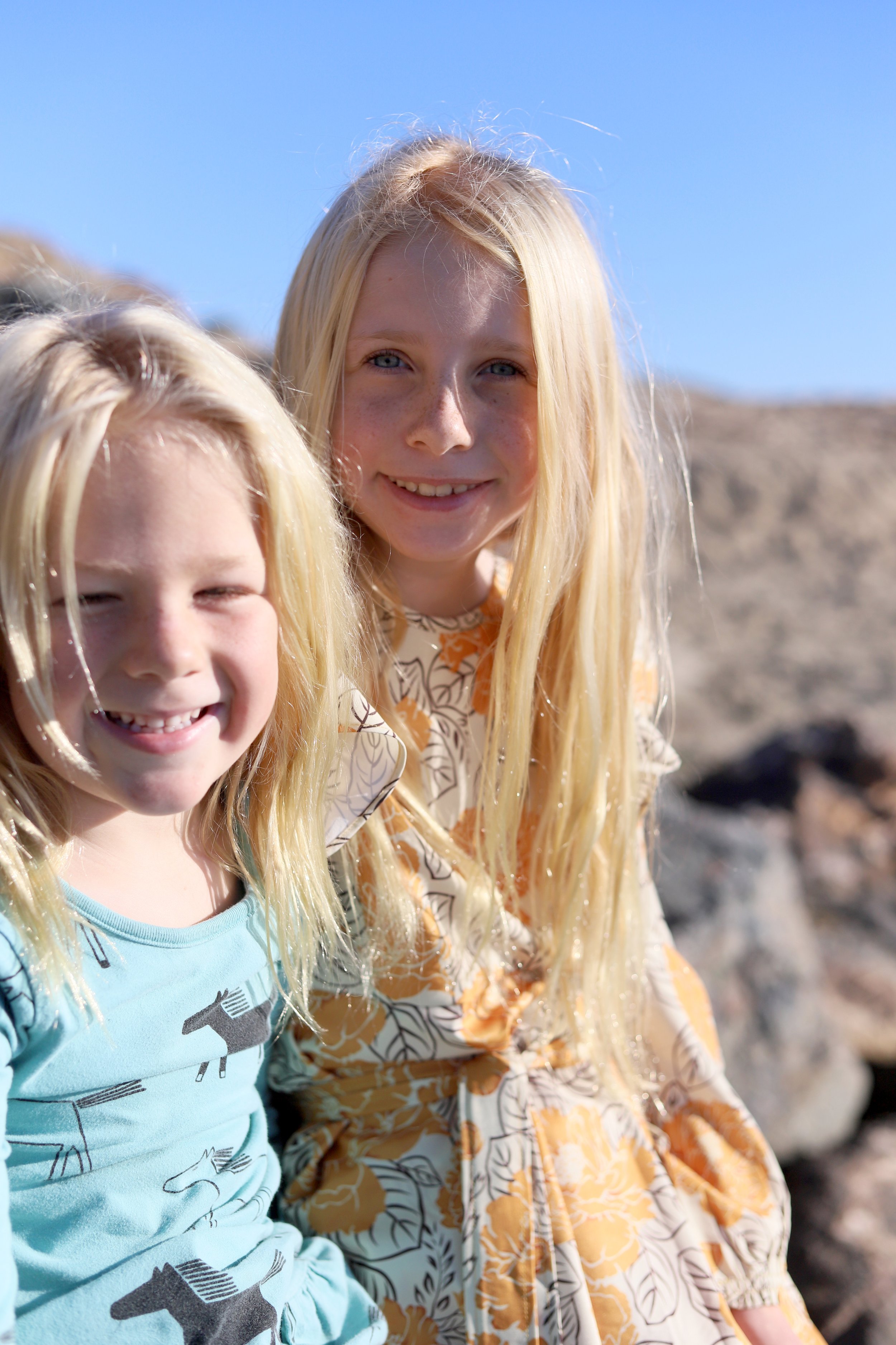 Two smiling girls with long blonde hair outdoors, with a clear blue sky and a rocky beach in the background.