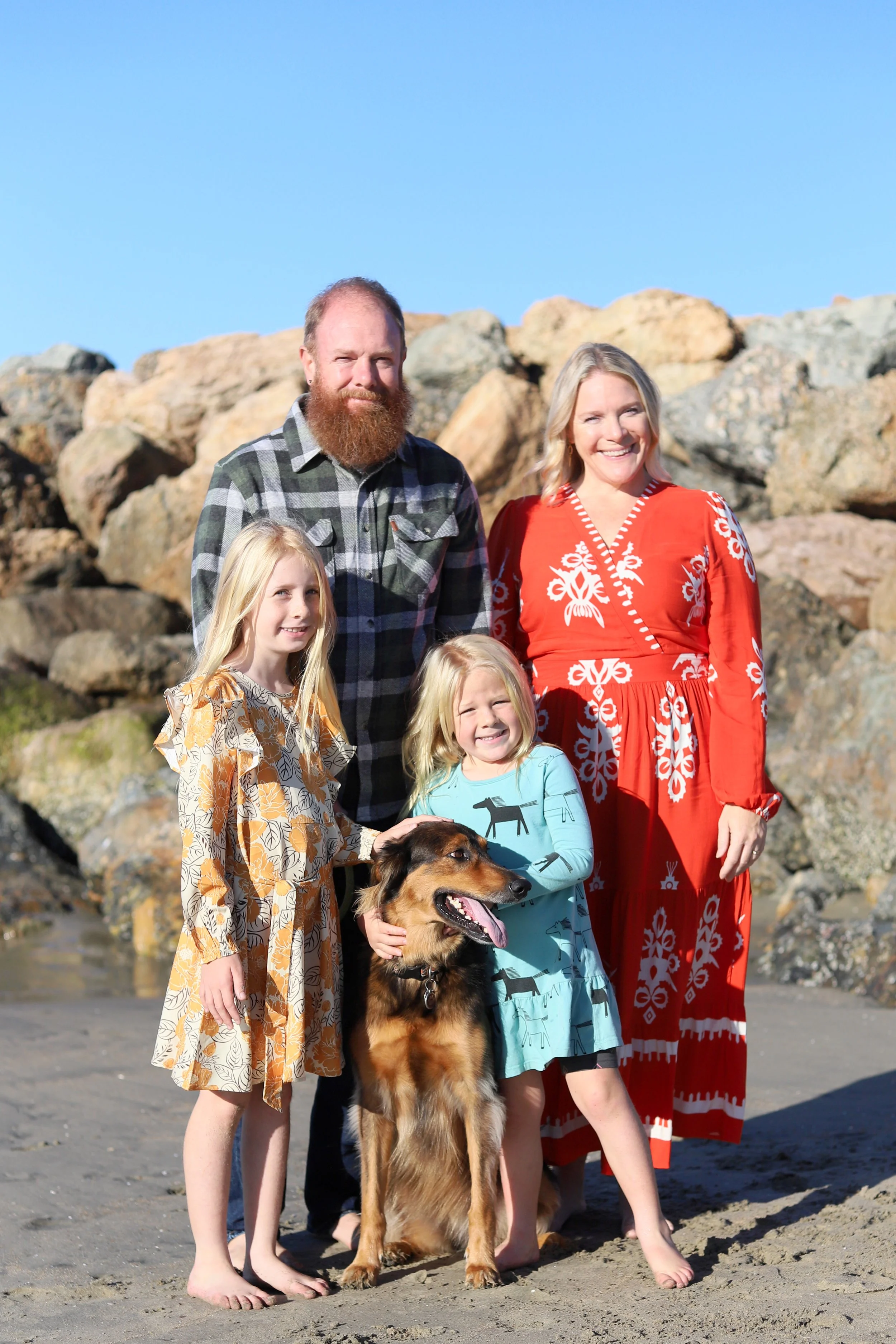 A family of five, including two children, a man, a woman, and a dog, posing on a beach with rocks in the background on a sunny day.