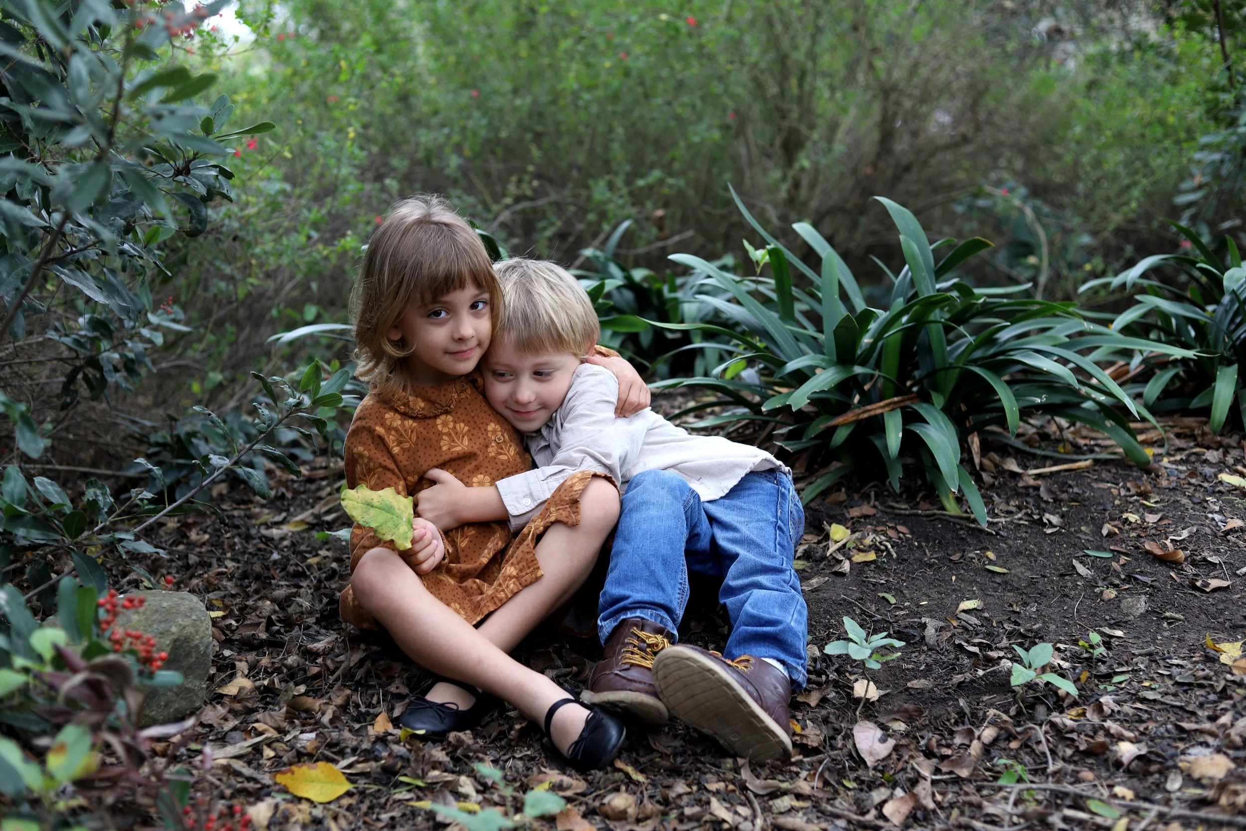 A girl and a boy sitting on the ground in a forest, hugging each other.