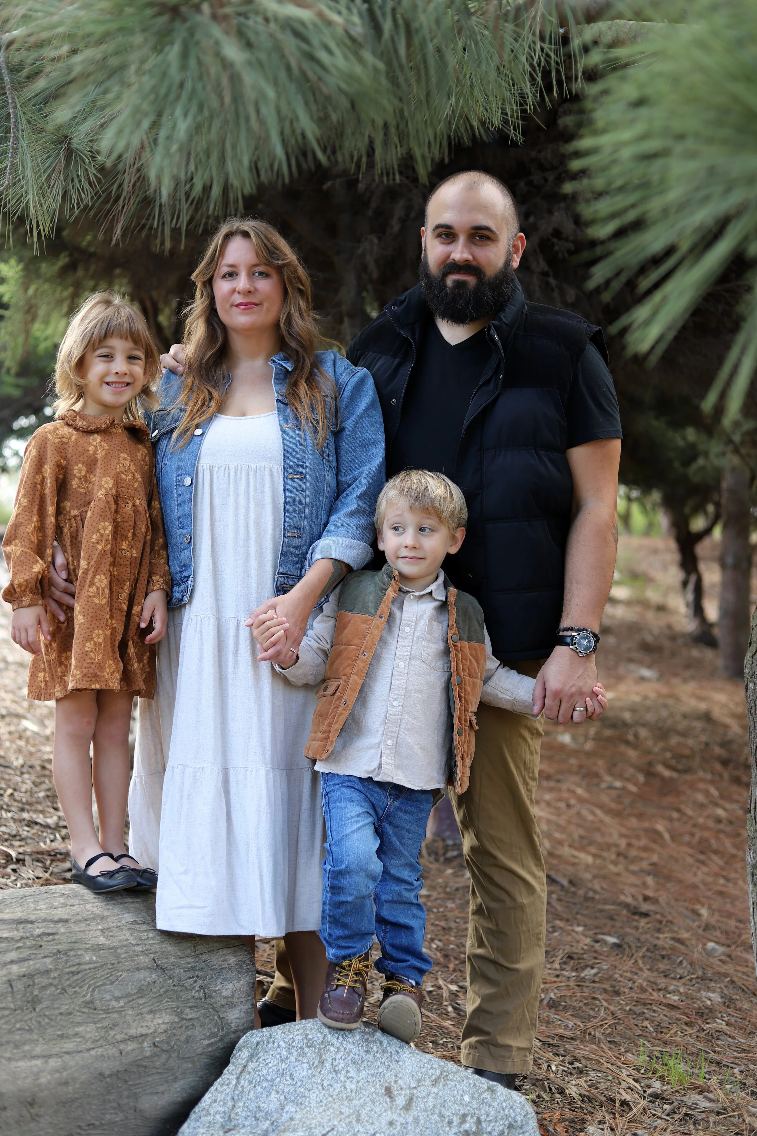 A family of four standing outdoors in a wooded area, holding hands and posing for a photo.
