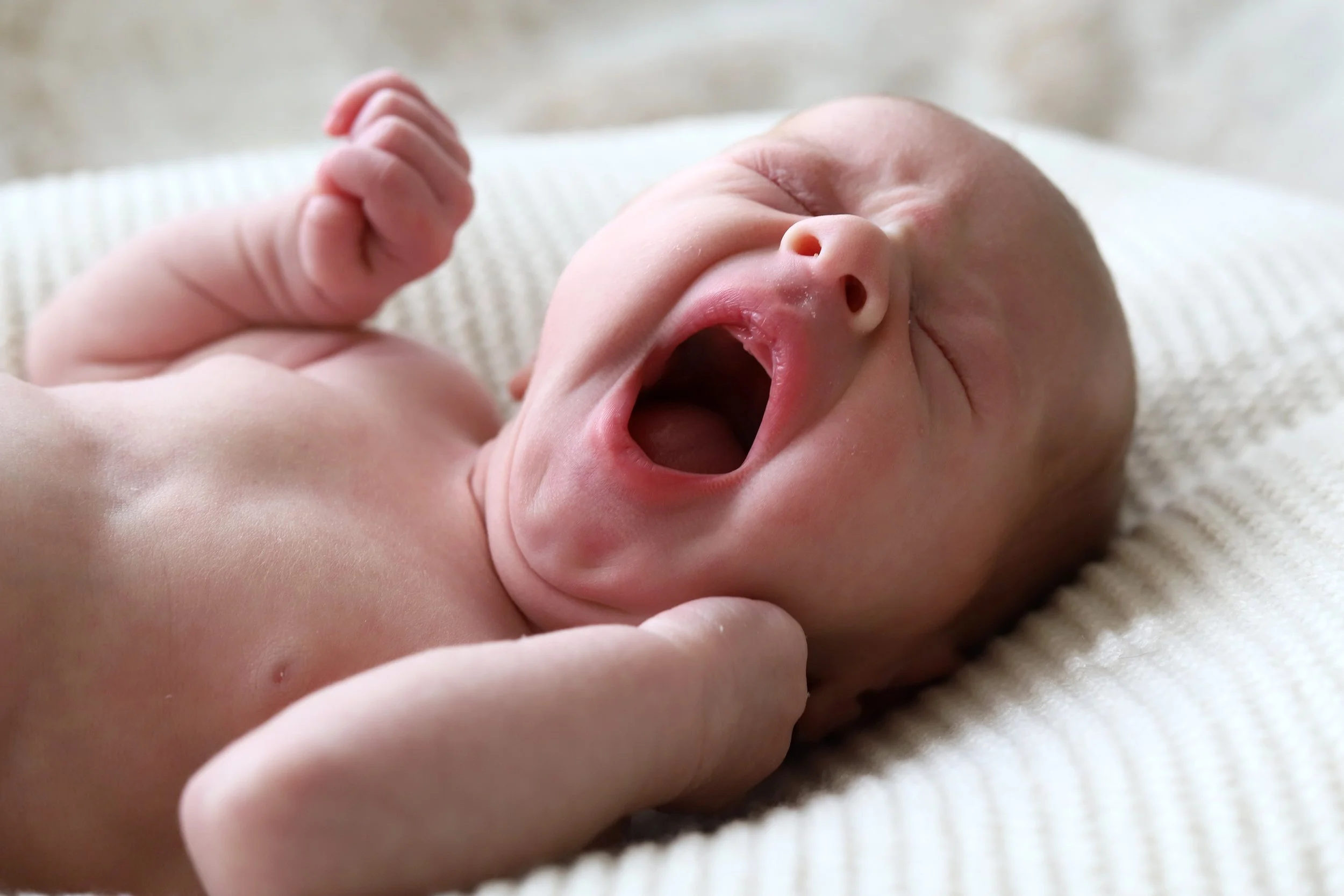 A close-up of a yawning baby lying on a textured white blanket, with a hand gently touching the baby's face.