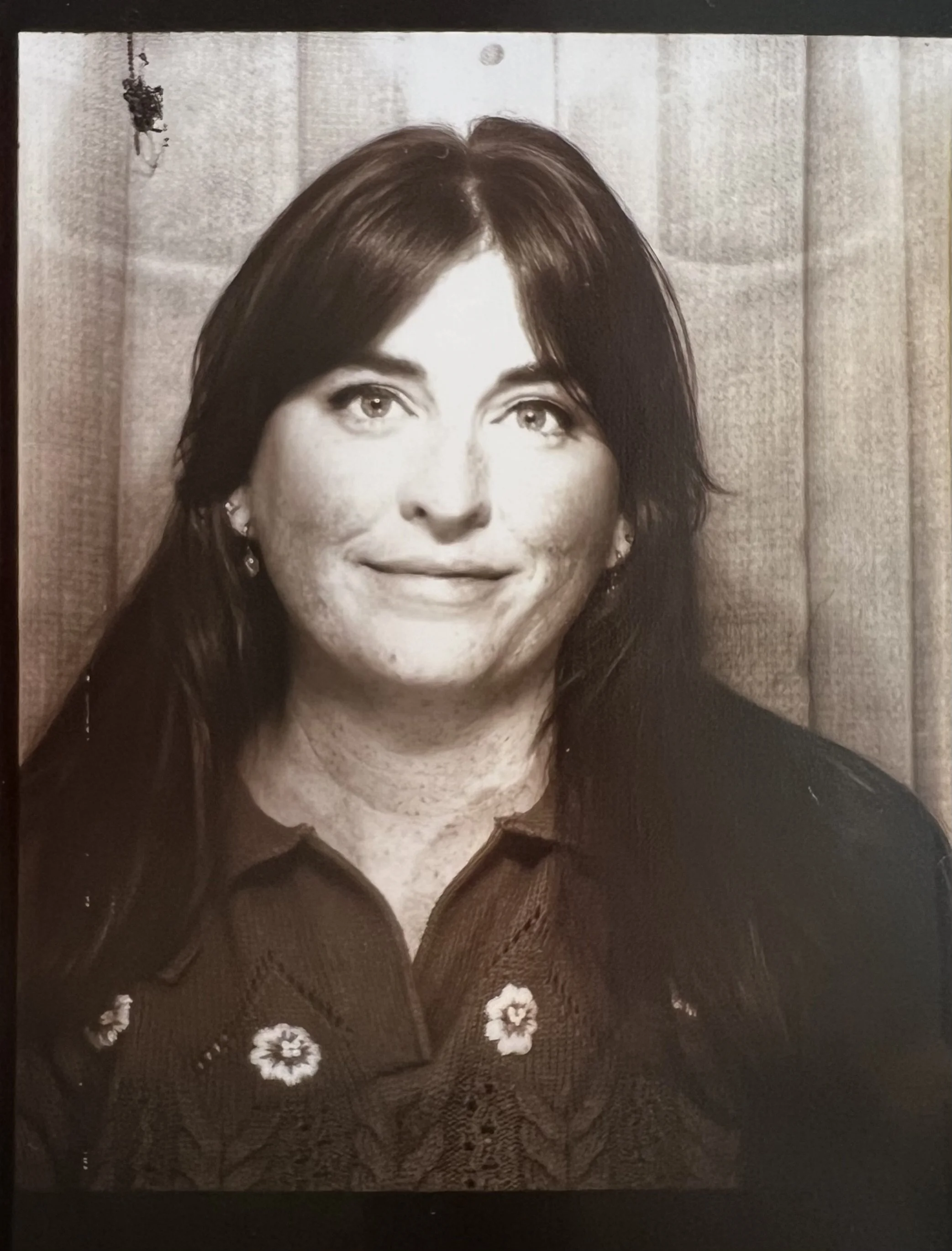 Black and white portrait of a woman with dark hair, smiling slightly, wearing earrings and a textured collared shirt with floral embroidery.