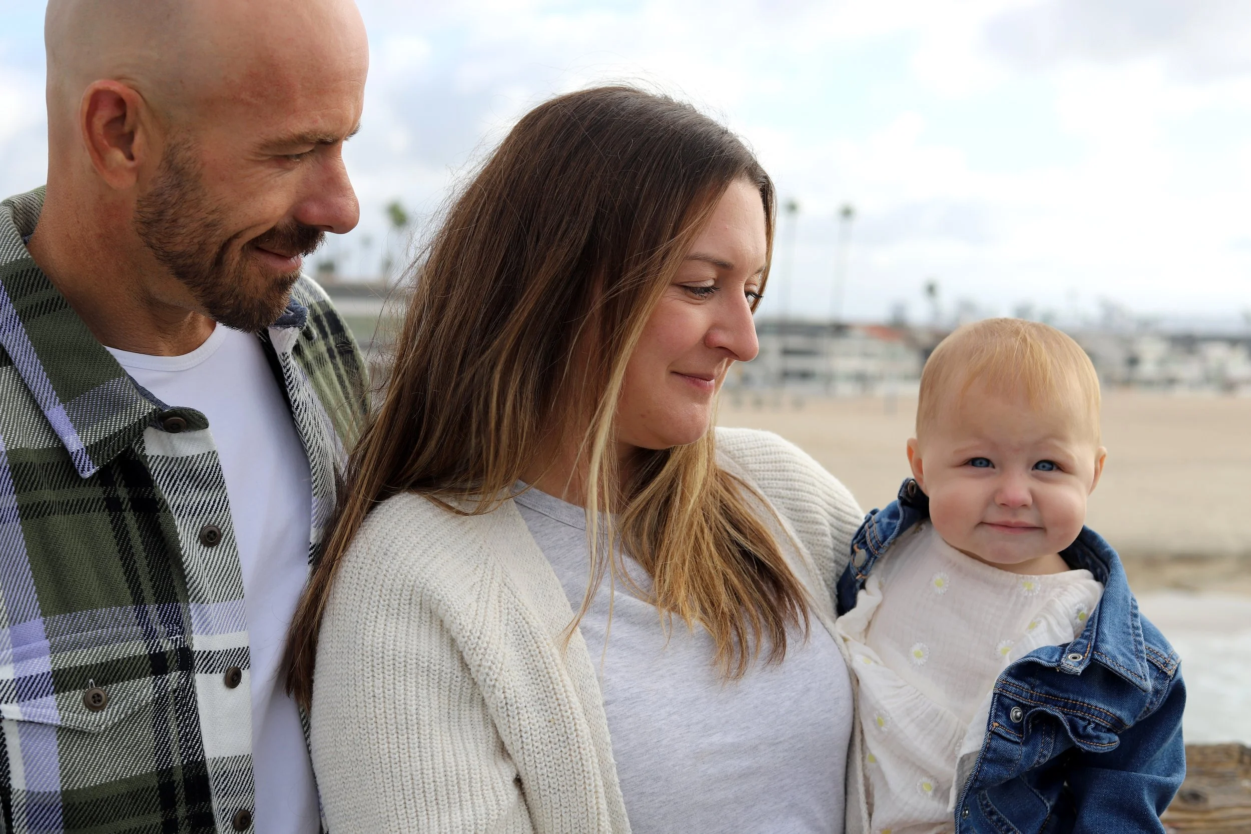 A family outdoors at the beach, with a man, a woman, and a young child, all smiling and looking at the camera.
