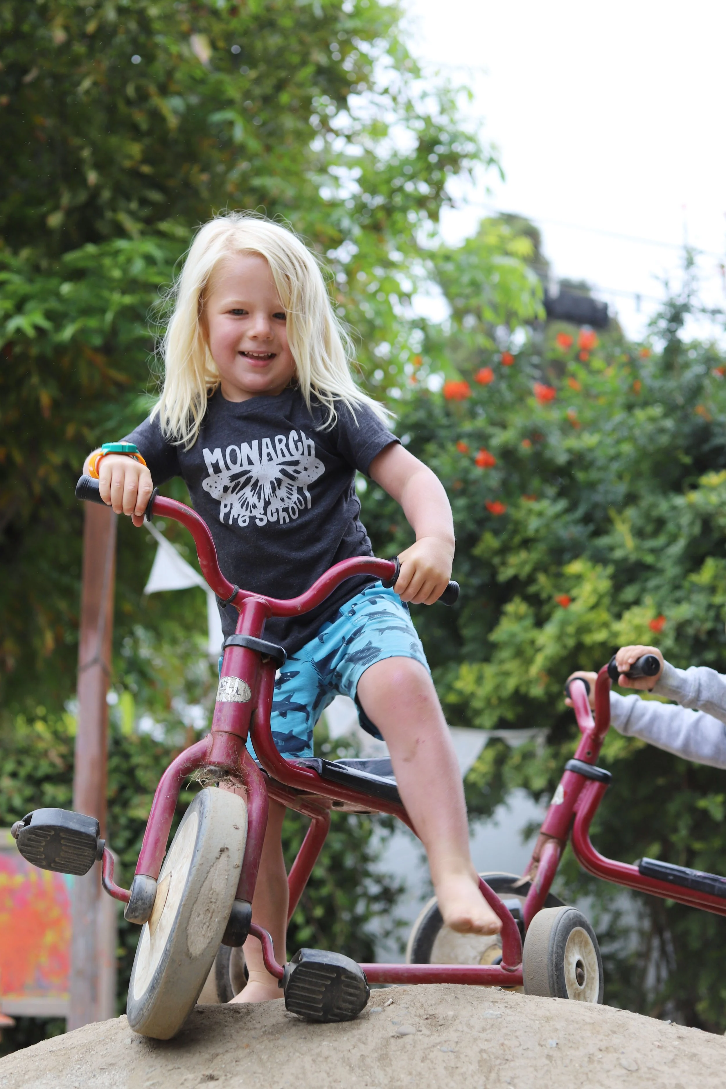 A young boy with blonde hair riding a tricycle on a dirt mound outdoors, smiling with greenery and trees in the background.
