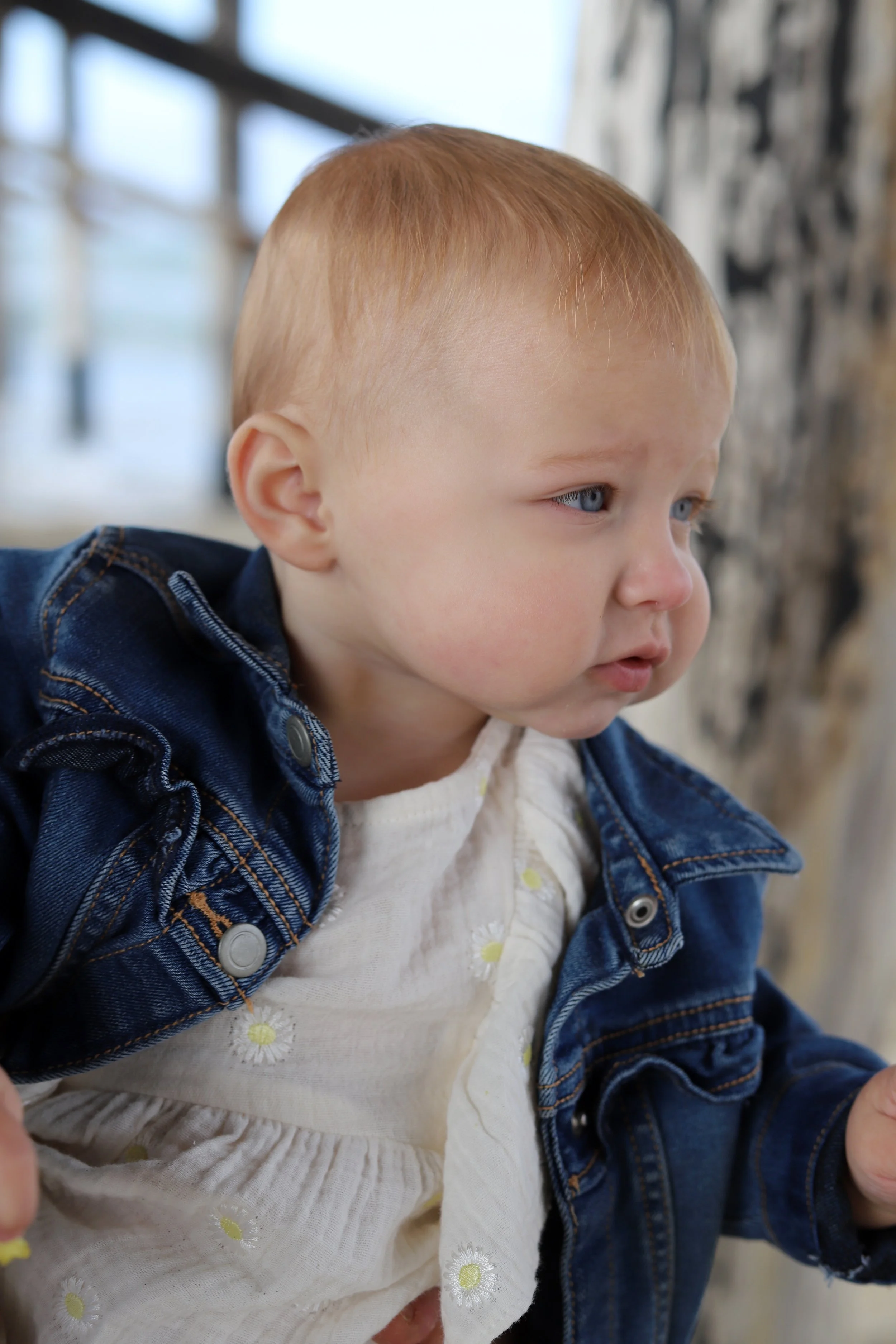 Close-up of a young child with red hair and blue eyes, wearing a denim jacket and a white dress with yellow flowers, looking away with a curious expression.