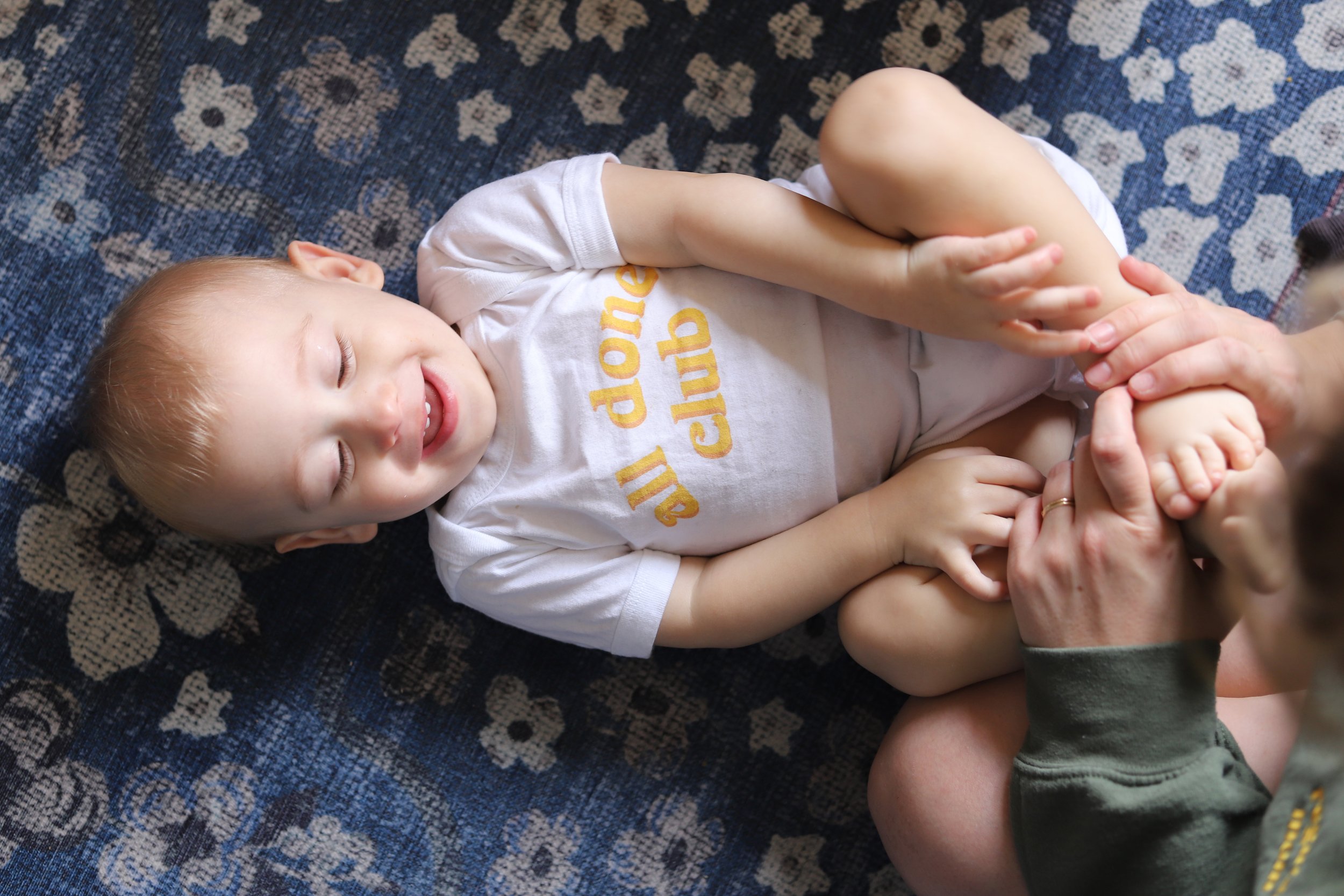 A young boy lying on his back on a patterned rug, smiling with his eyes closed. He is wearing a white t-shirt with yellow lettering and has light blonde hair. An adult's hands are holding his legs.