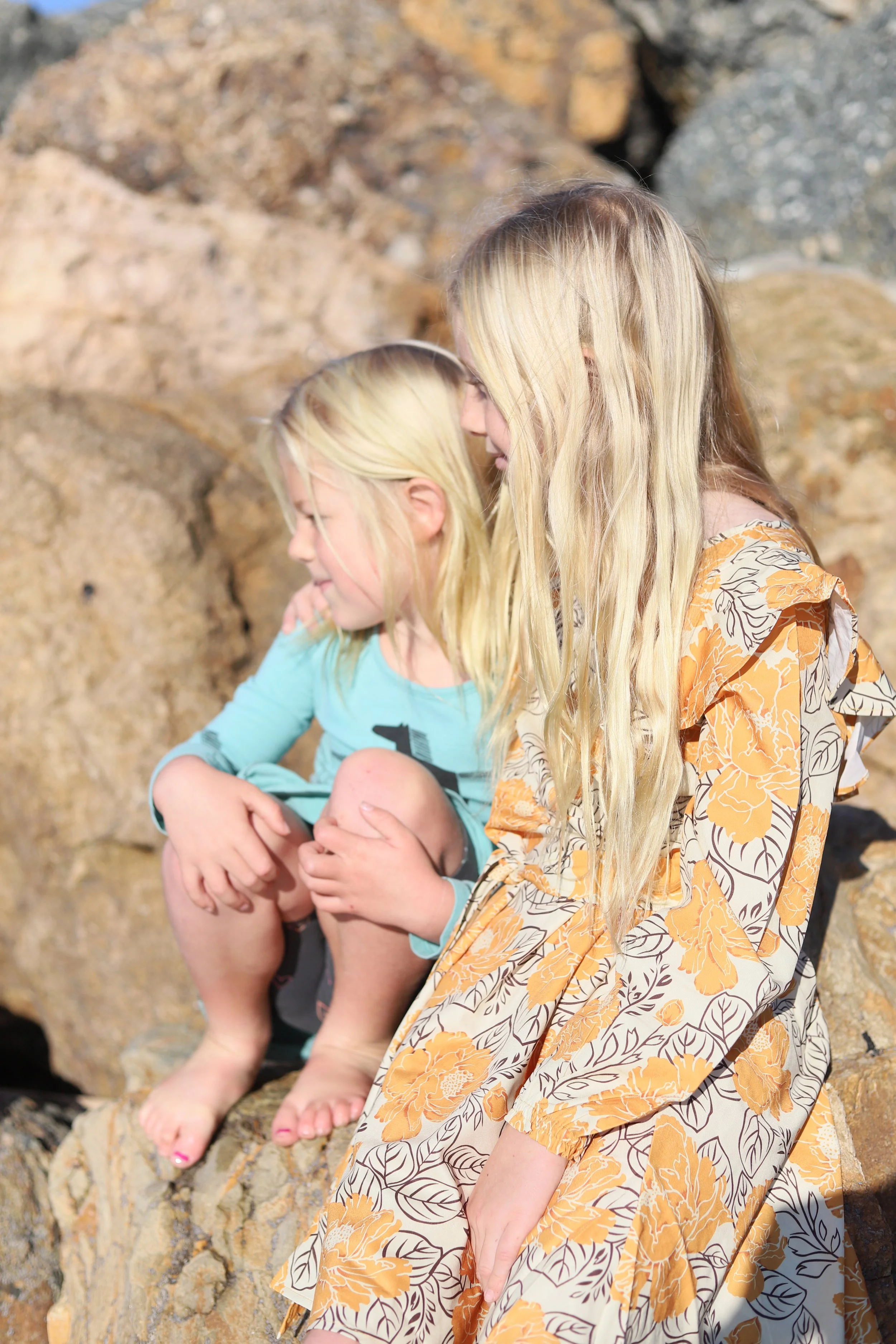 Two young girls with blonde hair sitting on rocks outdoors.