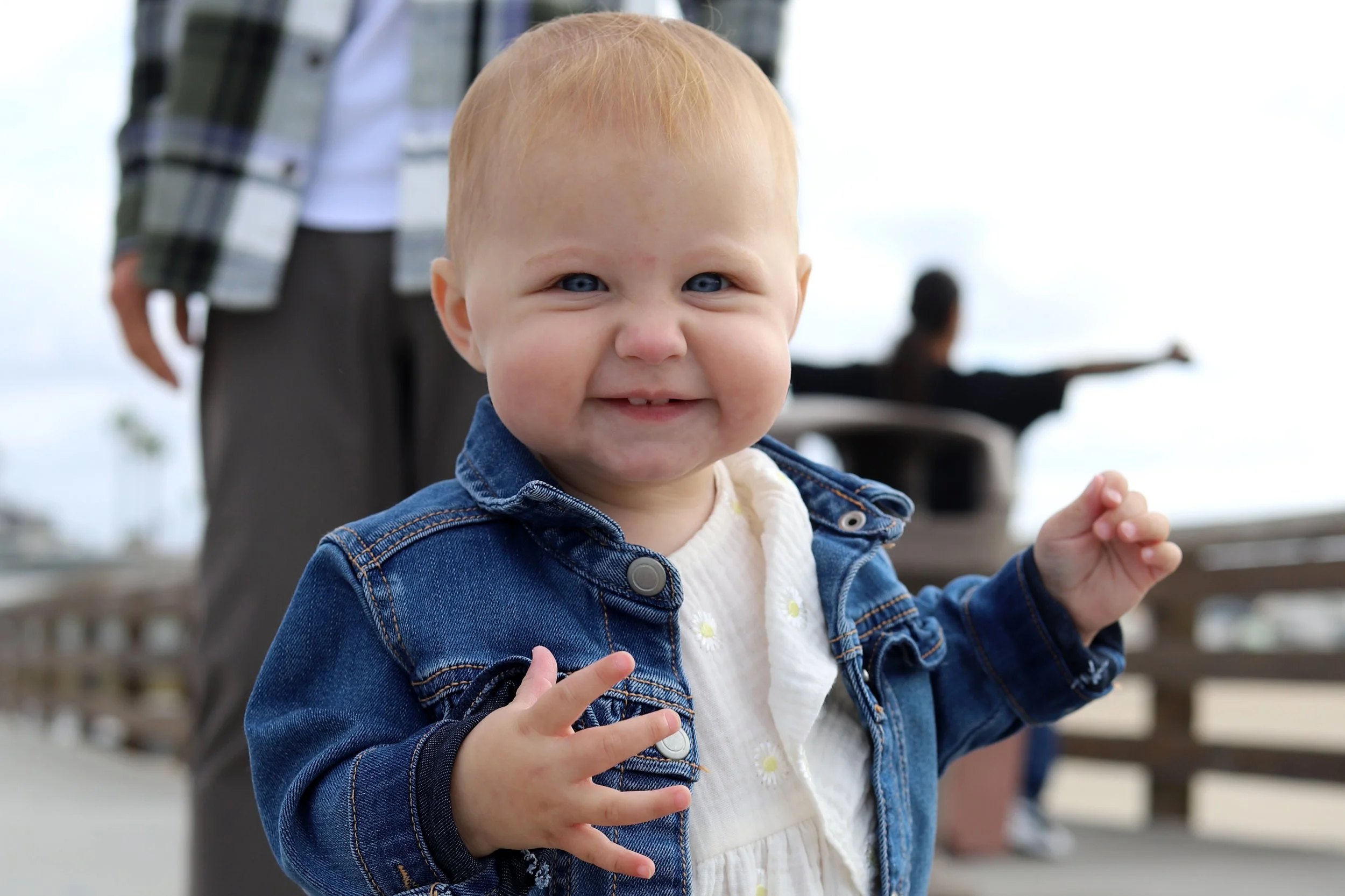 A happy toddler with blue eyes and red hair wearing a denim jacket and a white dress with daisy embroidery, standing outdoors by a wooden fence with a blurred person in the background pointing with her arm outstretched.