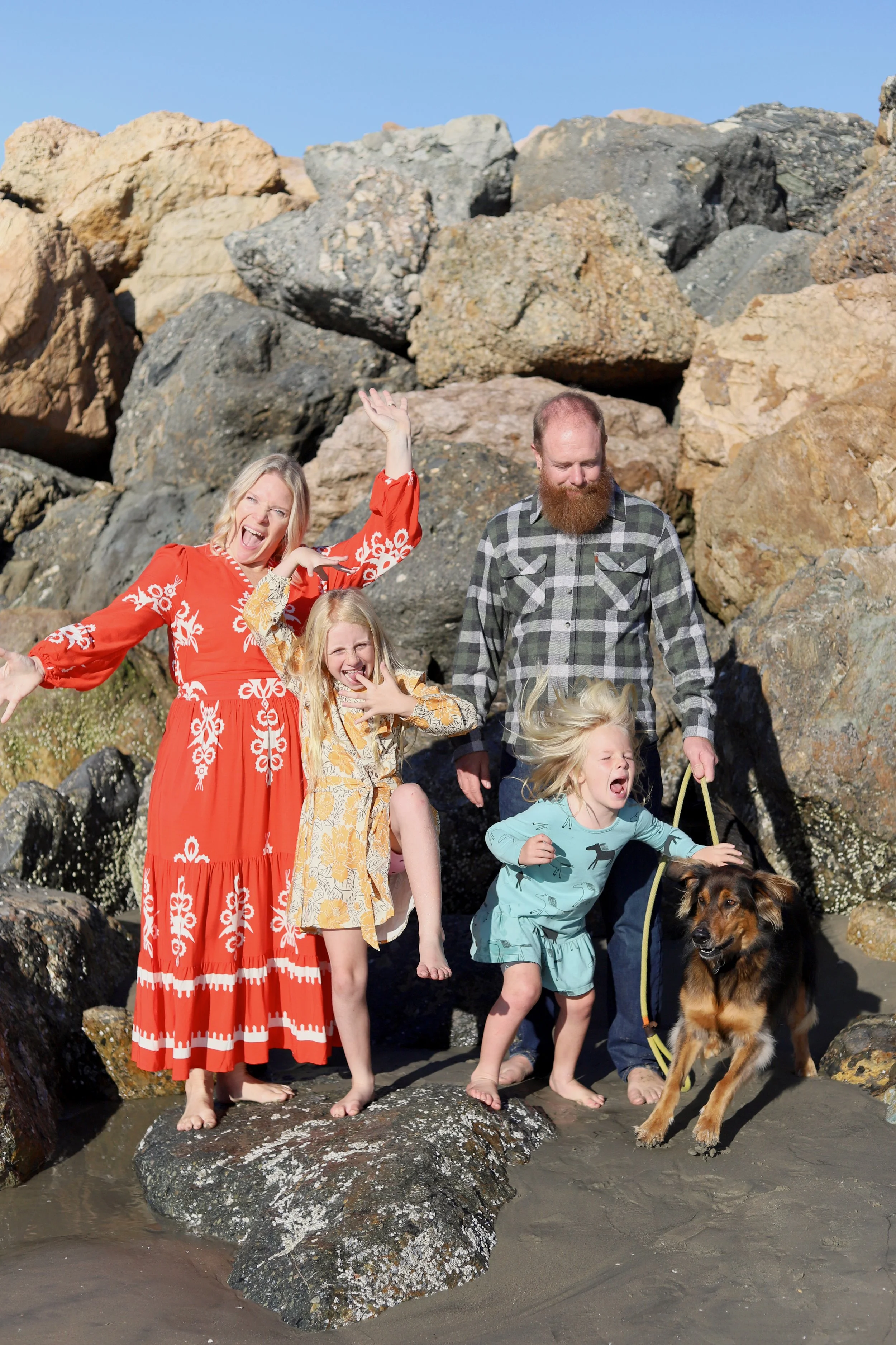 A family of five, including two women, two young girls, and a man, with a dog, standing on rocks at a beach with a background of large rocks and clear blue sky. The women and the girls are making playful, expressive faces, and the man is looking down