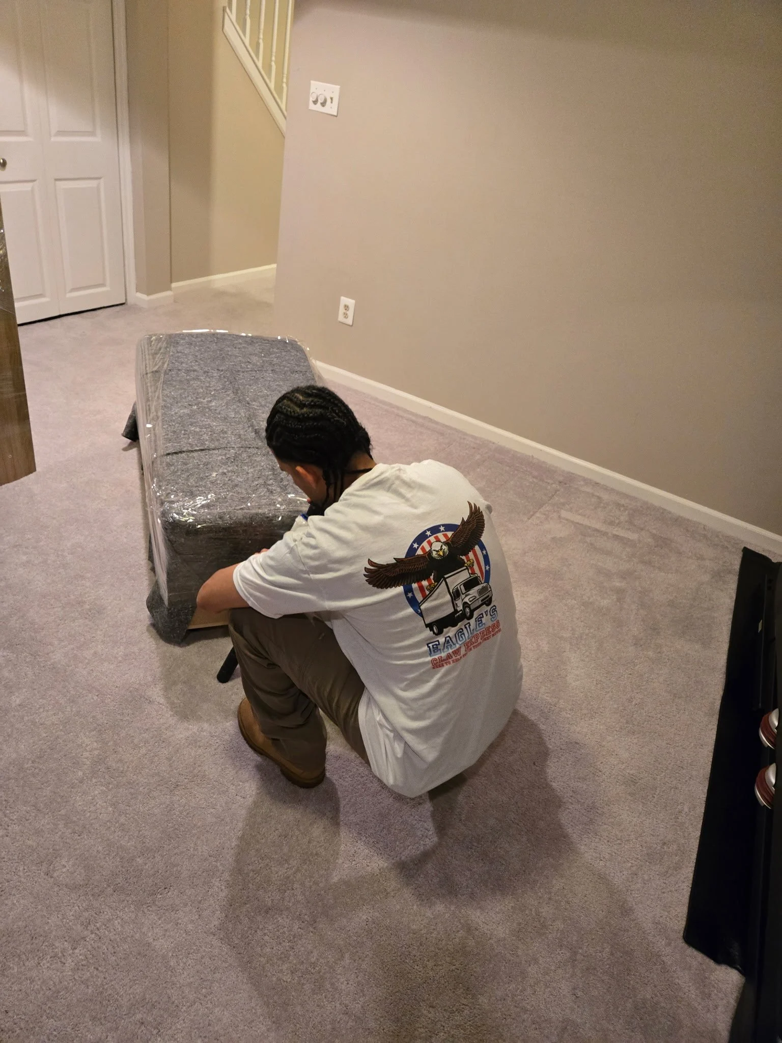 Person kneeling on the carpeted floor, unpacking a large wrapped object, in a room with beige walls and carpet, near a white door and electrical outlets.