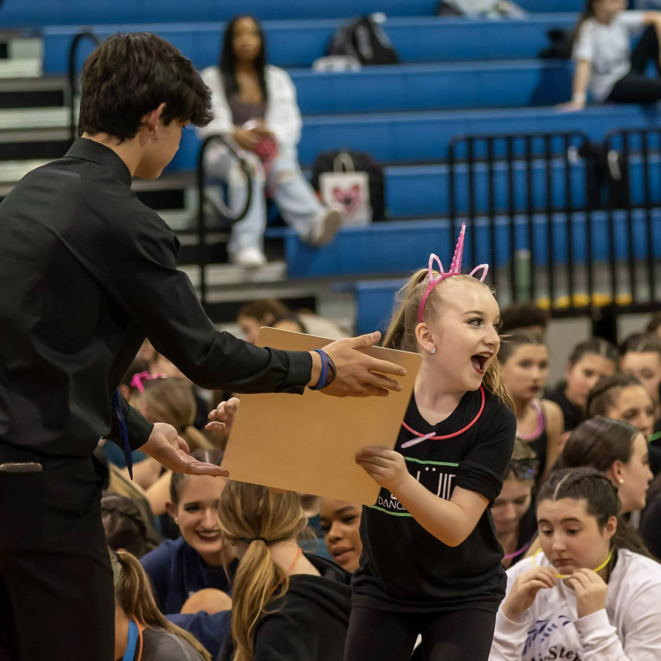 A young girl at a dance competition receives an award from a man, wearing a pink unicorn headband, while a crowd of girls sits on the floor in the background.