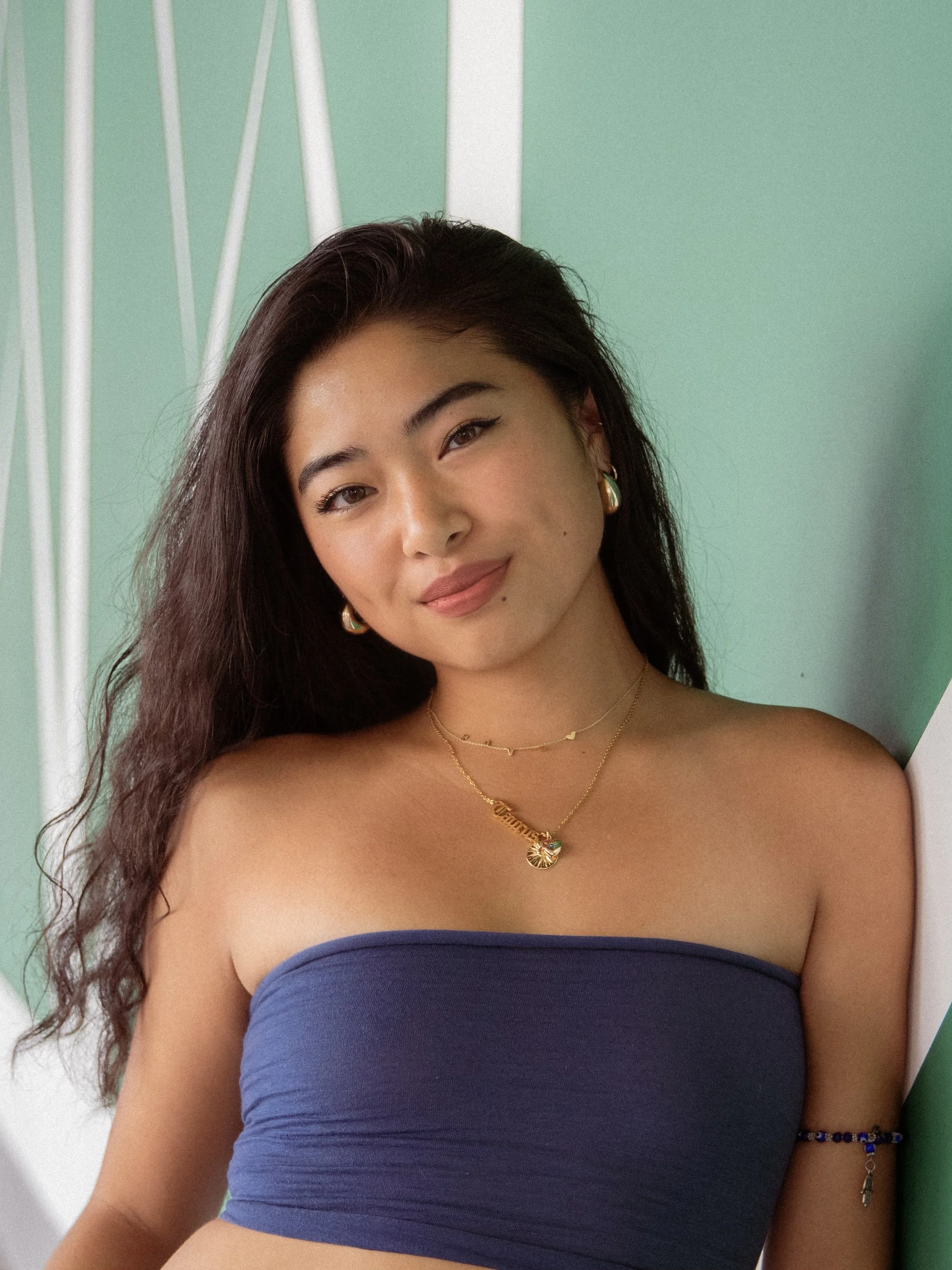 A young woman with long dark hair and light makeup, wearing gold jewelry including earrings and layered necklaces, is smiling softly while leaning against a green wall with white geometric decor.