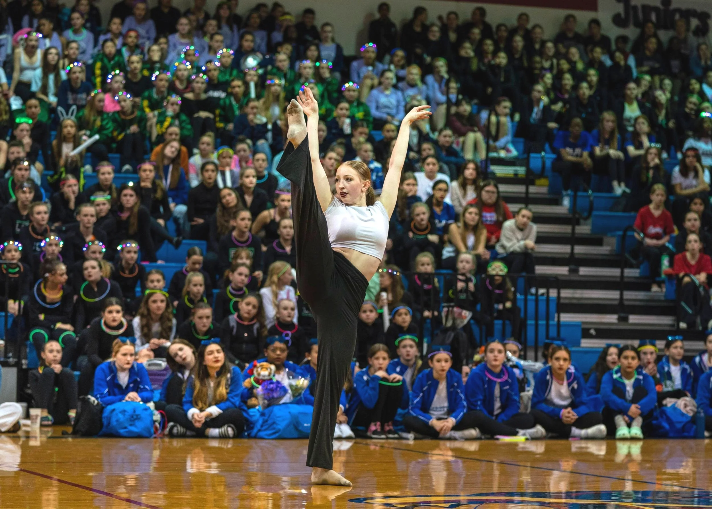 A young woman performing a dance move on a gymnasium floor in front of a large audience of children and teenagers seated on bleachers.