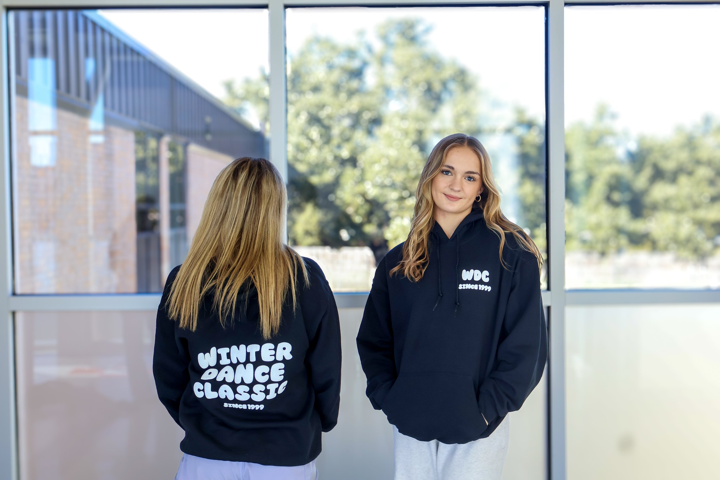 Two young women wearing black hoodies with white text; one faces away with her hoodie reading "Winter Dance Classic," the other smiles toward the camera with her hoodie reading "WDC Since 1999"; they stand inside near a window with an outdoor scene of trees and buildings visible.
