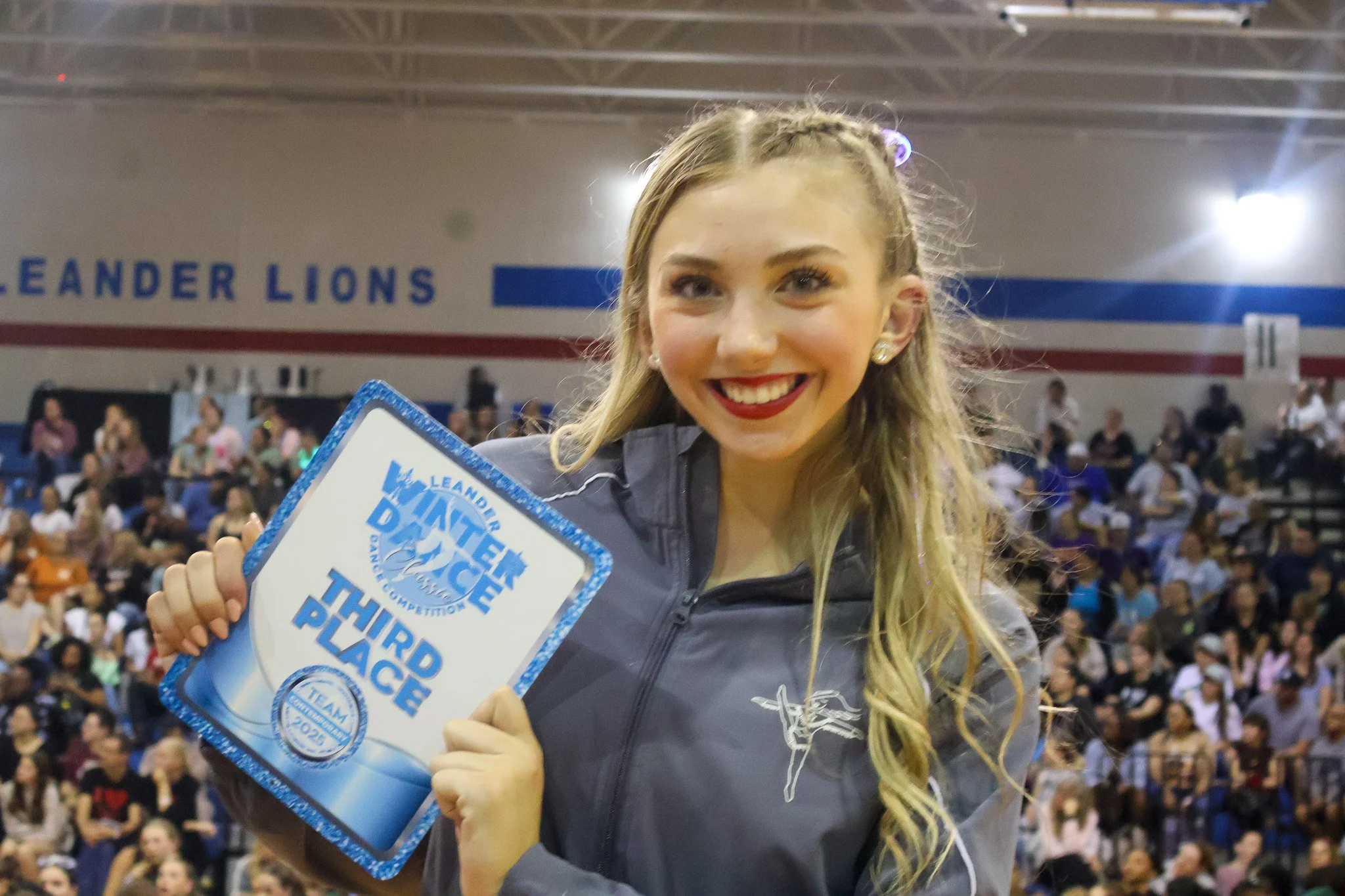 A young woman at WDC competition holding a third place award in front of a crowd in an indoor gymnasium. The background has a banner that reads 'LEANDER LIONS' and the woman is smiling.