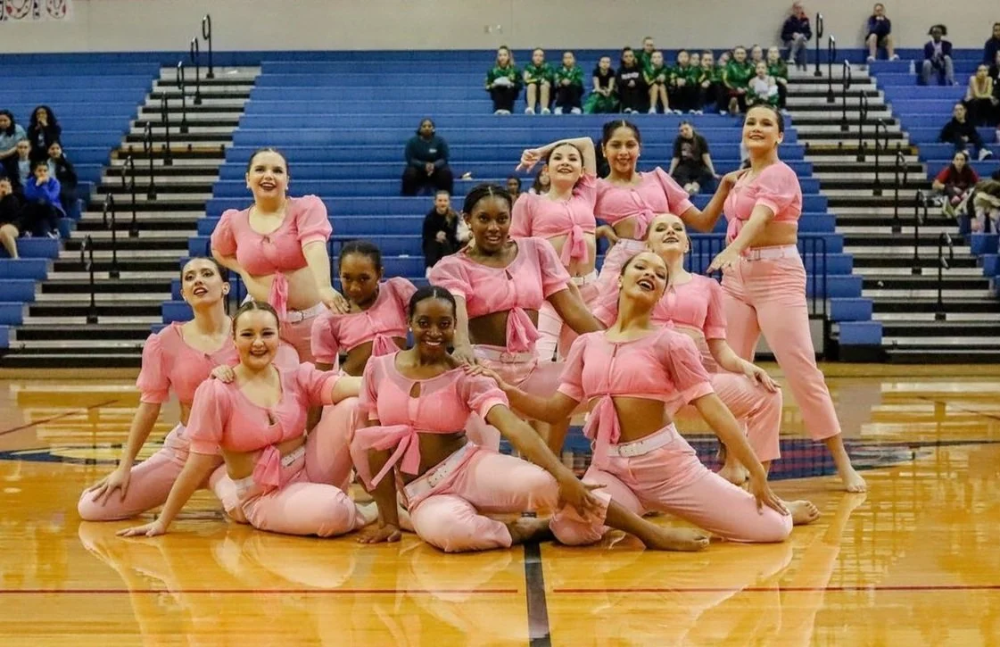 A group of young female cheerleaders in pink outfits posing for a photo on a gymnasium floor with bleachers and spectators in the background, at WDC Dance Competition in Leander, TX. The premier Central Texas dance competition and contest.