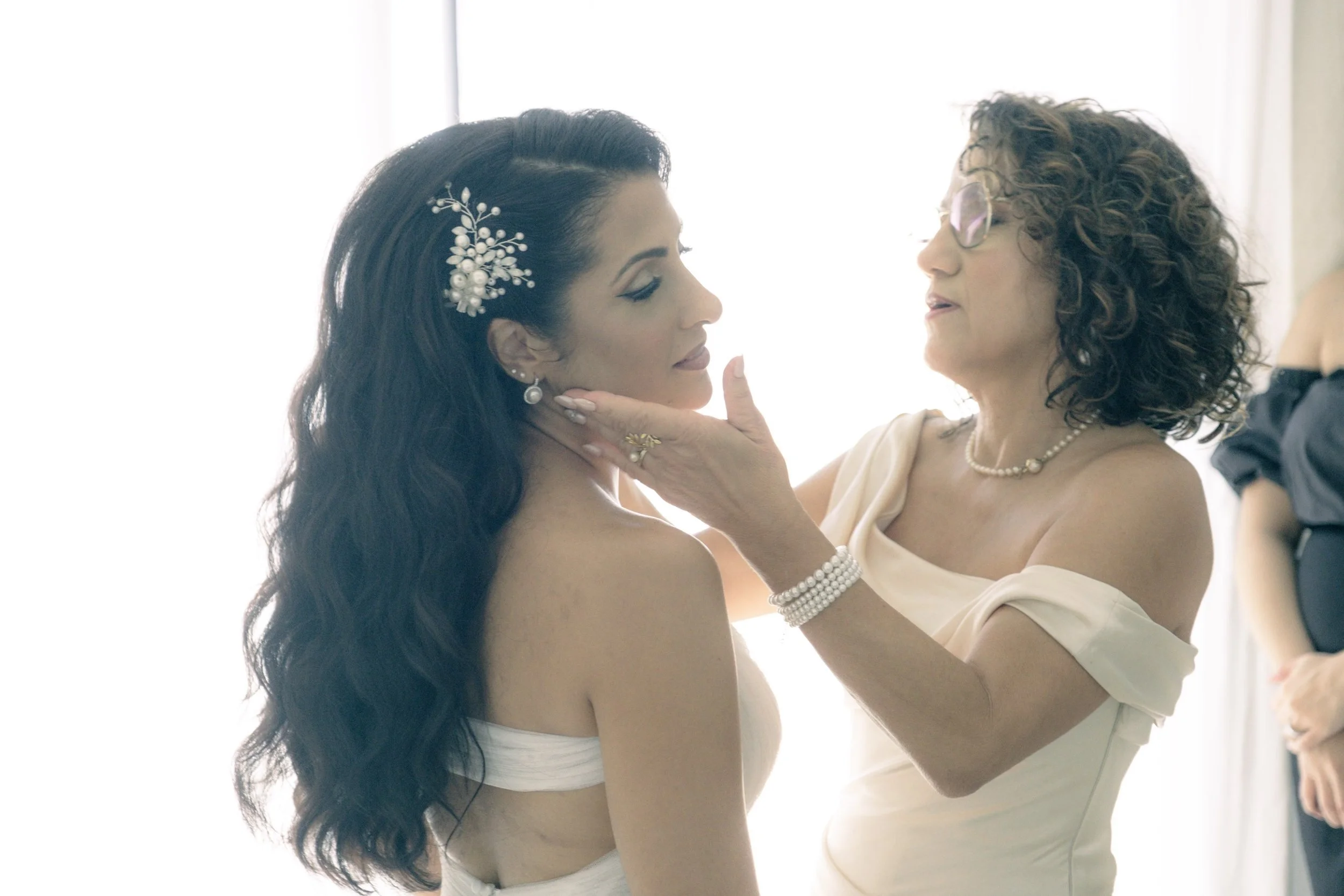 Une femme touche le visage d'une jeune femme qui porte une robe de mariée, dans une pièce lumineuse.