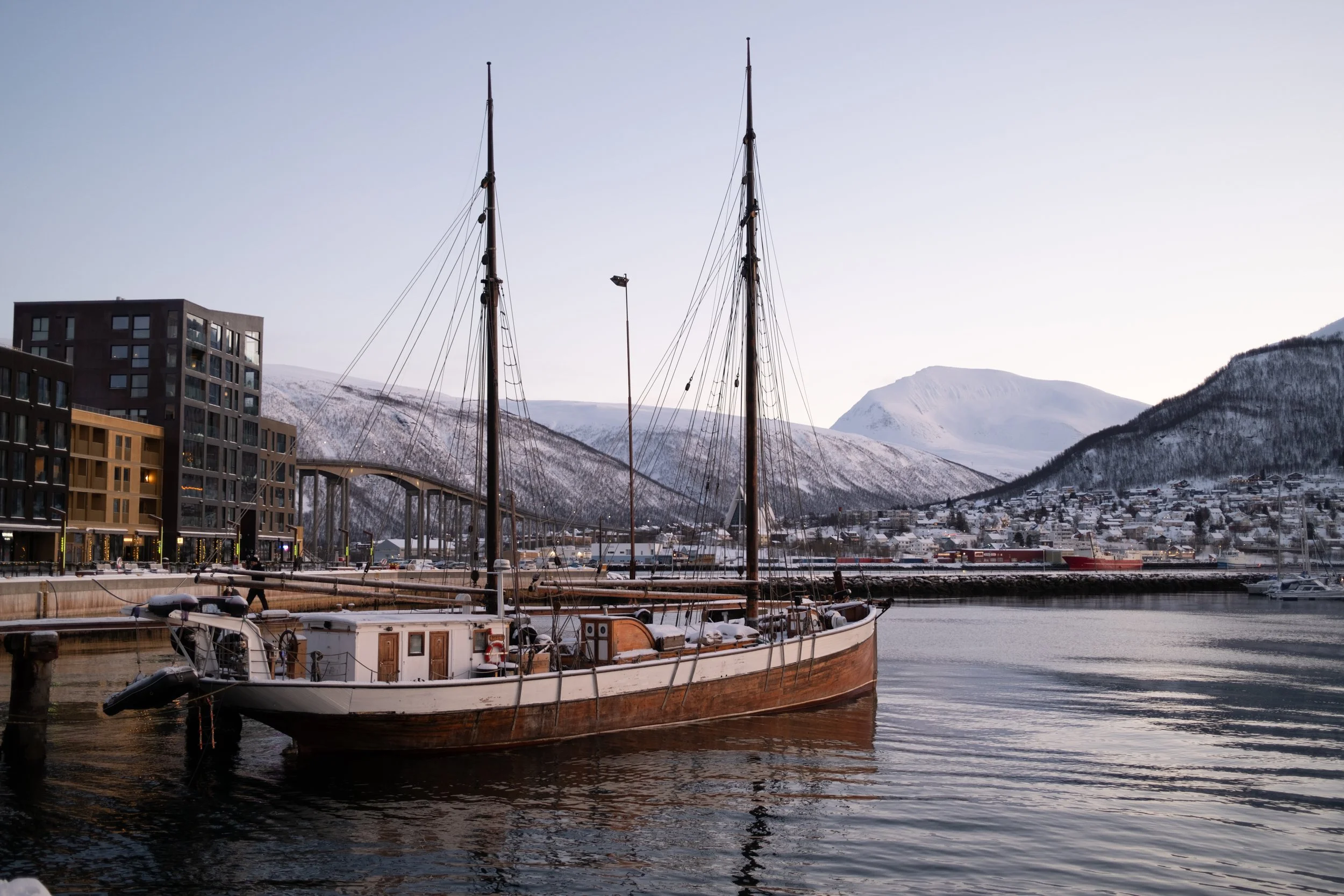 Tromsø, Norvège. Un voilier en bois amarré dans un port, avec des montagnes enneigées en arrière-plan, des bâtiments modernes et un pont dans une ville côtière. - Photographe voyage à La Réunion