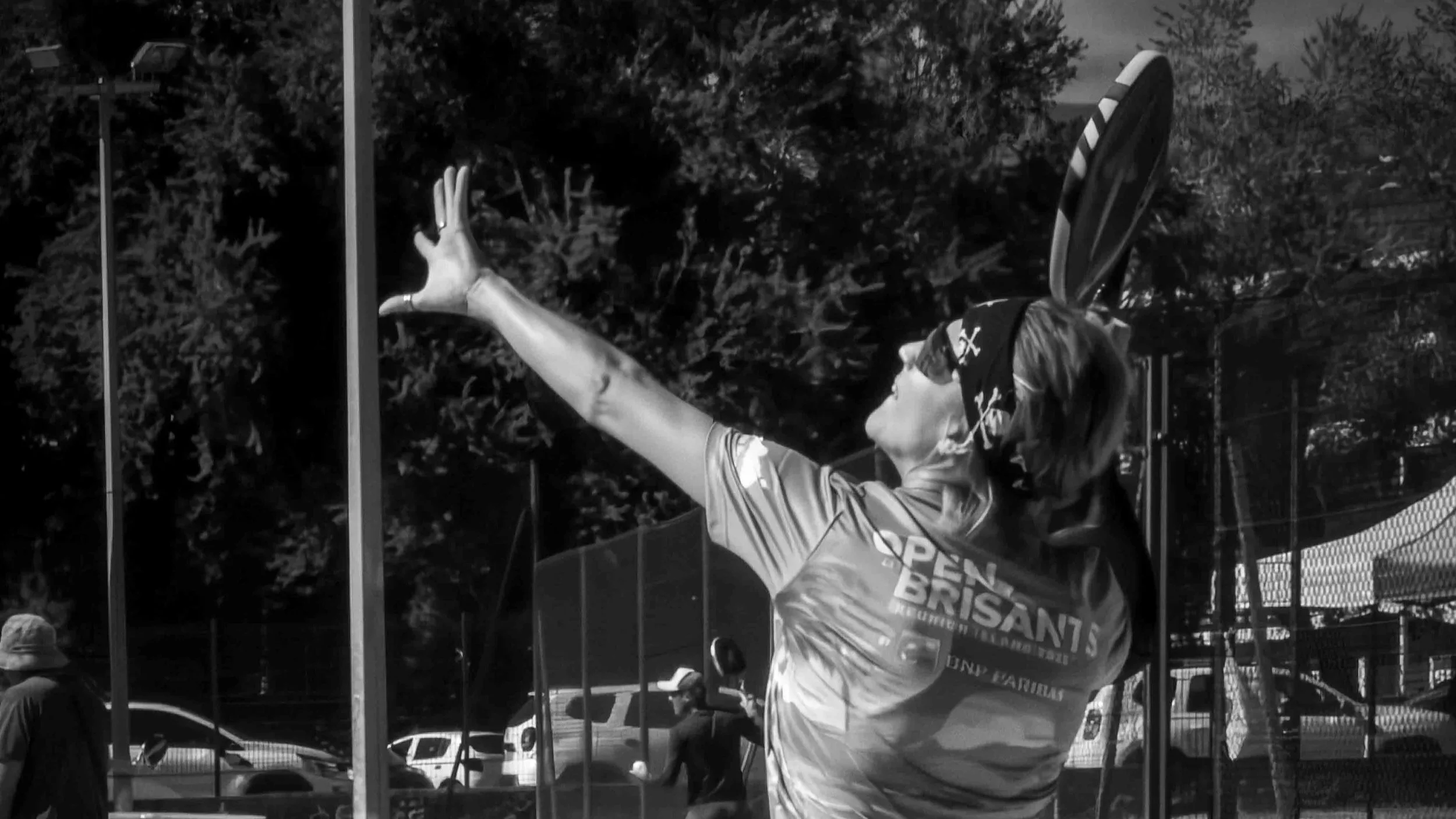 Une femme jouant au Beach paddle tennis s'apprêtant à servir, porte un bandeau et une chemise avec le texte 'OPEN des BRISANTS' à l'arrière-plan un terrain de tennis entouré d'une clôture et de voitures - Photographe sportif à La Réunion