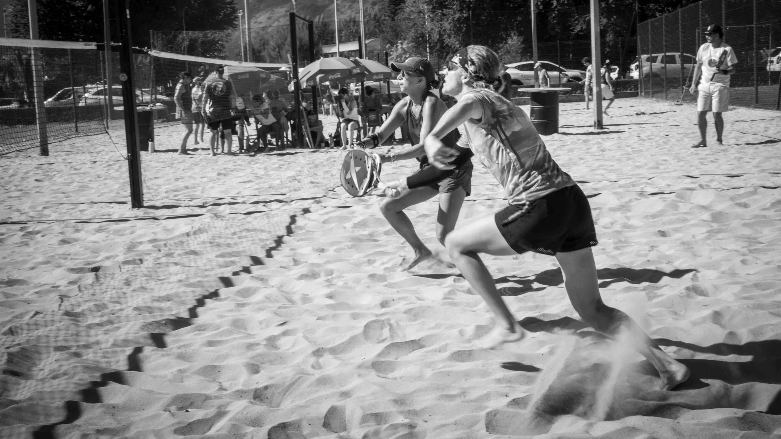 Deux femmes jouent au Beach paddle tennis sur le sable, vêtues de shorts et t-shirts, avec un filet de volley-ball en face. En arrière-plan, des spectateurs et d'autres personnes près de voitures et d'une tente sous un parasol. - Photographe sportif 