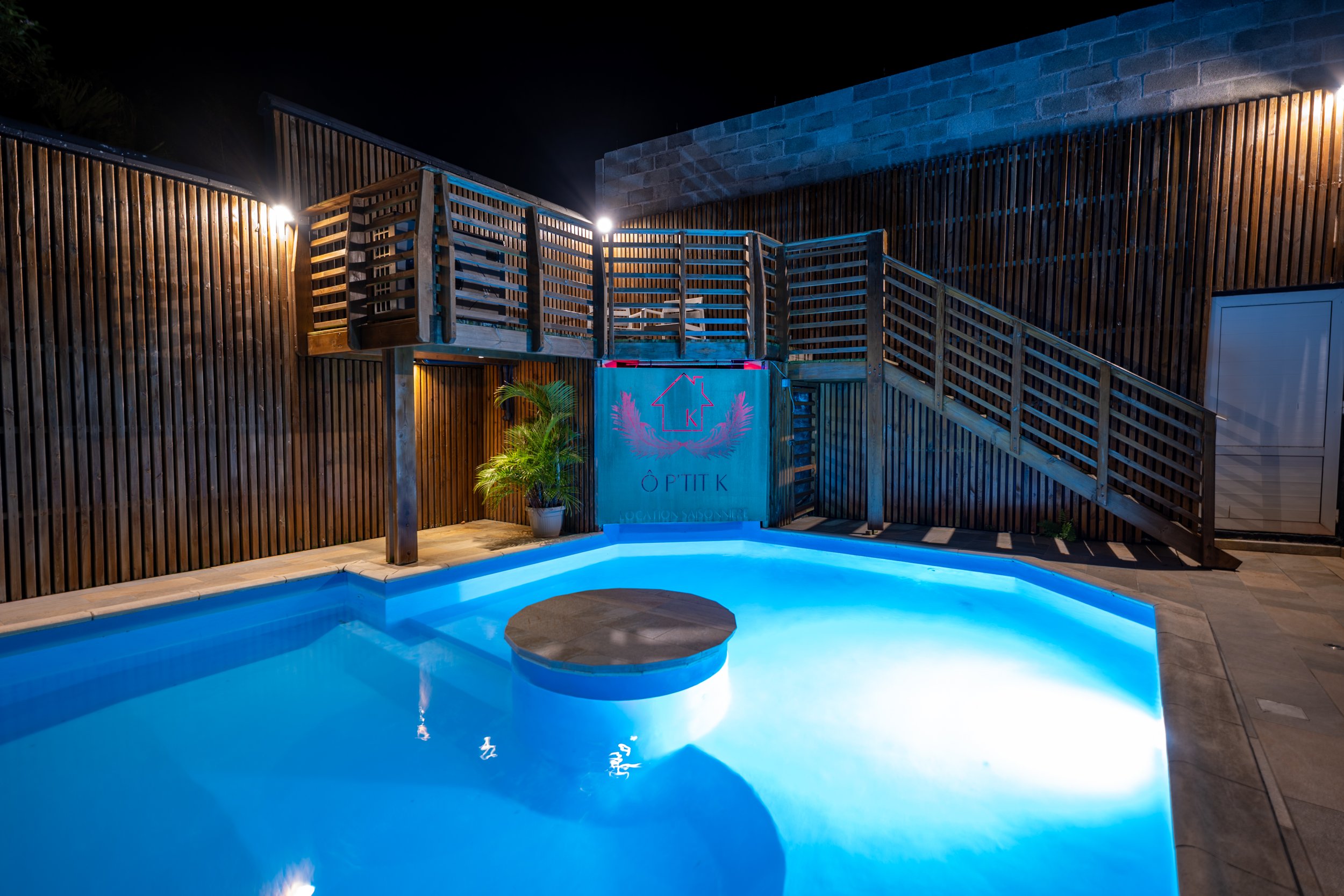 Piscine éclairée la nuit avec un mur en bois, un escalier en bois et une plante en pot - Photographe immobilier à La Réunion