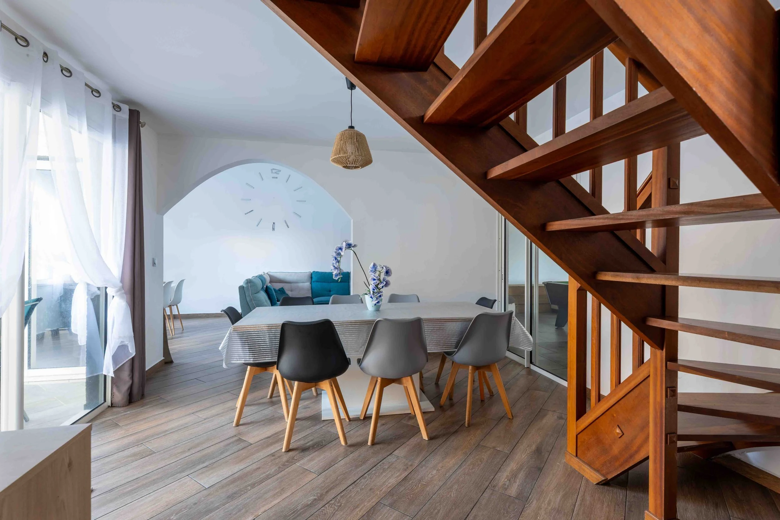Intérieur moderne d'une maison avec une cuisine ouverte, une table à manger entourée de sept chaises modernes, un escalier en bois non fini et un salon au fond avec un canapé bleu, Photographe immobilier à La Réunion
