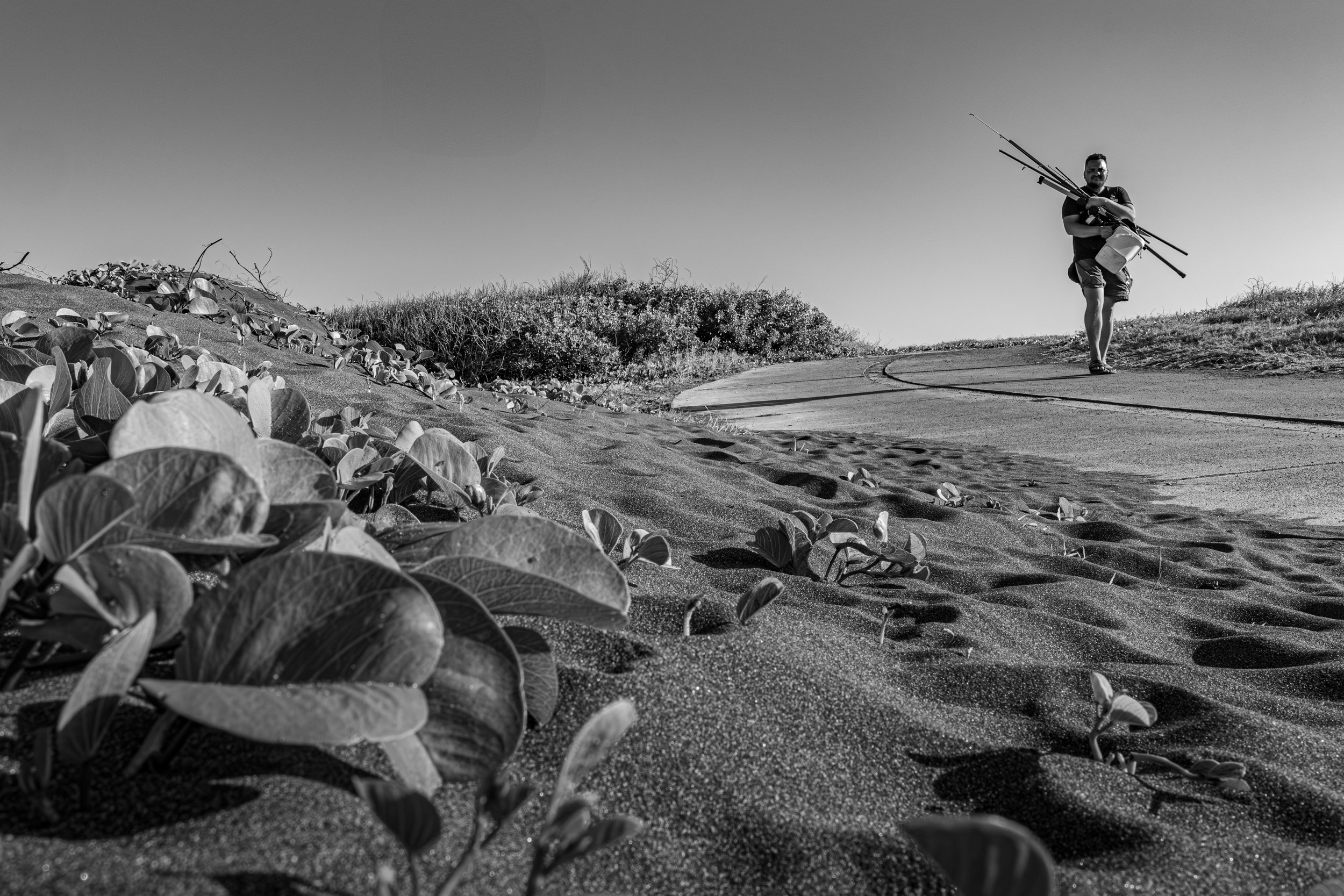 Scène en noir et blanc d’un pêcheur marchant avec ses cannes sur une plage de sable à La Réunion — Photographe lifestyle