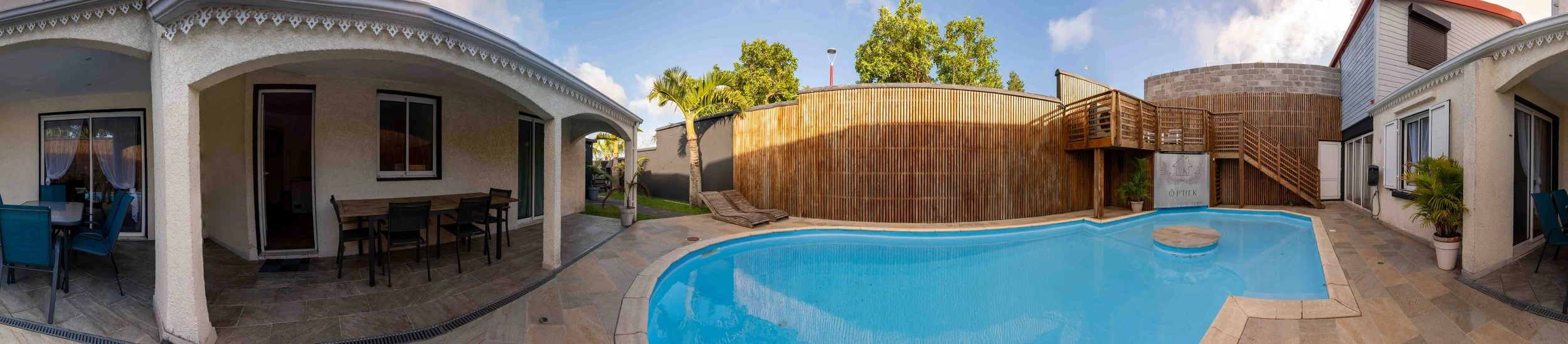 Photo d'une cour arrière avec piscine, terrasse, canapés, chaises, et petits arbres, entourée de murs en bois et en pierre, sous un ciel bleu avec quelques nuages.