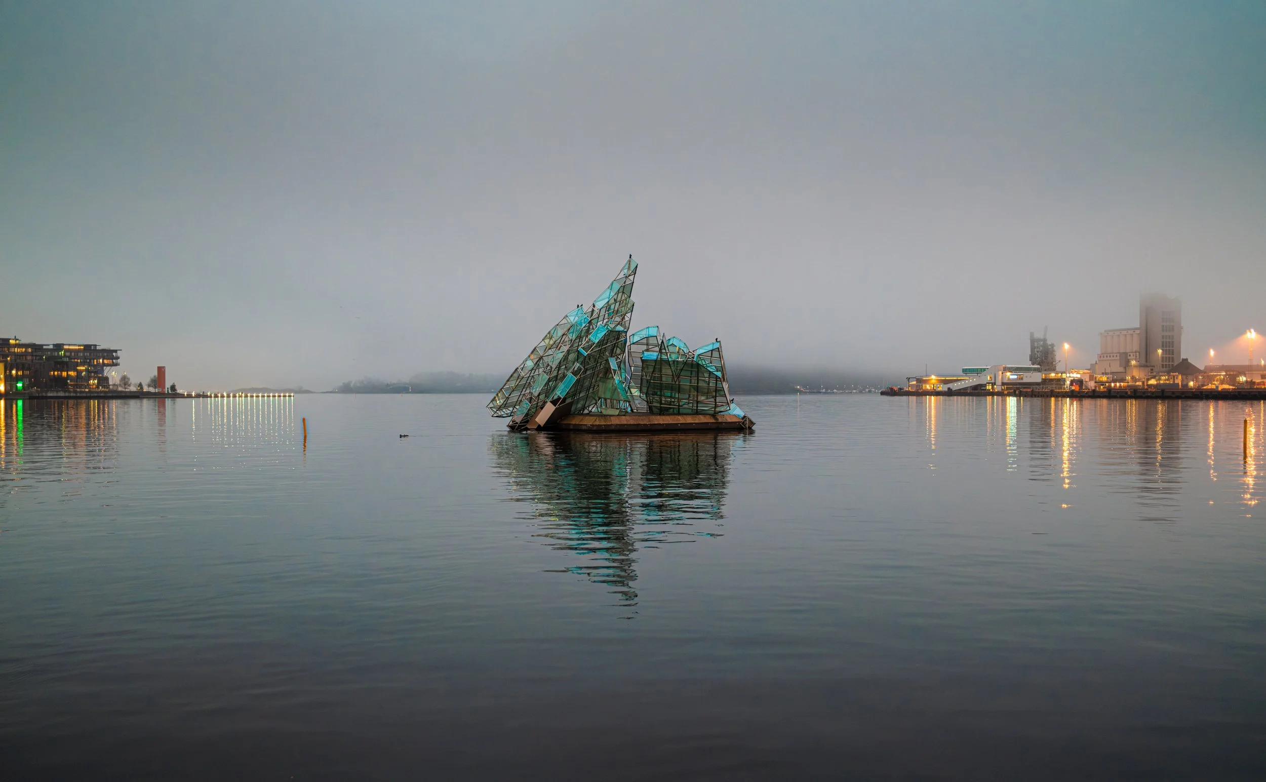 Oslo, Norvège. Une sculpture en forme de bâtiment de verre au centre d'un plan d'eau, avec une ville en arrière-plan, sous un ciel nuageux le soir. - Photographe voyage à La Réunion