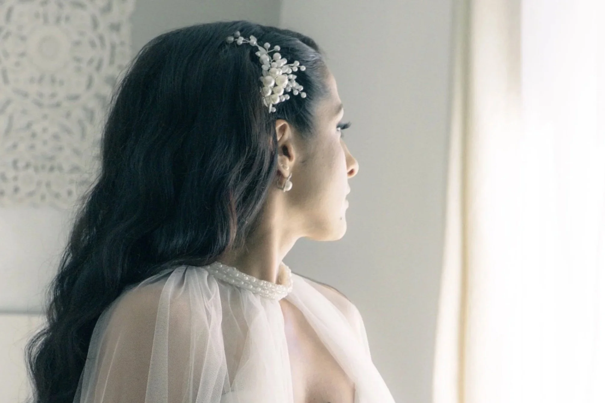 Mariée en robe blanche, regardant par la fenêtre, cheveux longs, coiffée d'une broche ornée de perles et d'ornements floraux blancs.