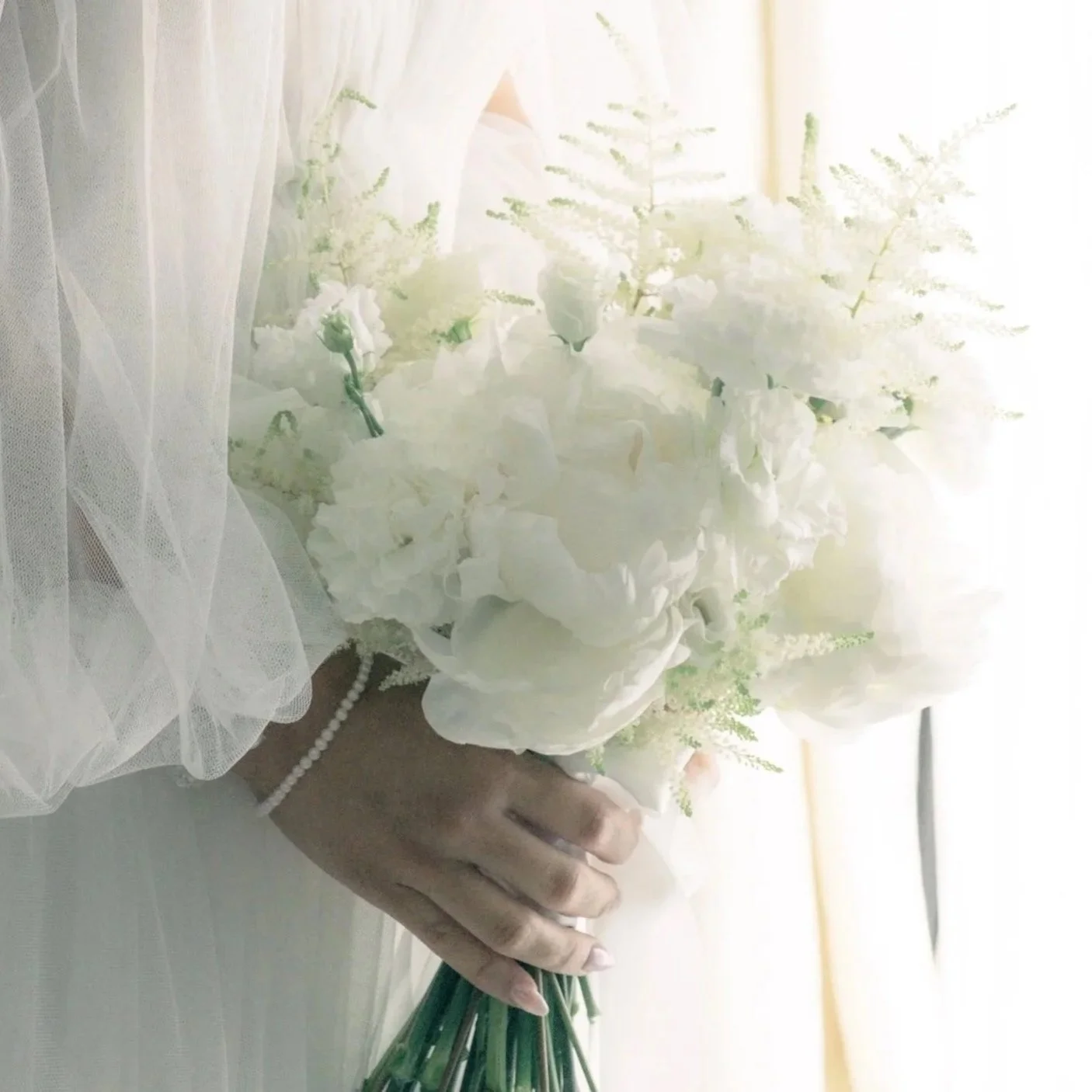 Main tenant un bouquet de fleurs blanches, probablement pour un mariage, avec une main portant un bracelet de perles blanc. Tissu léger et translucide en arrière-plan.
