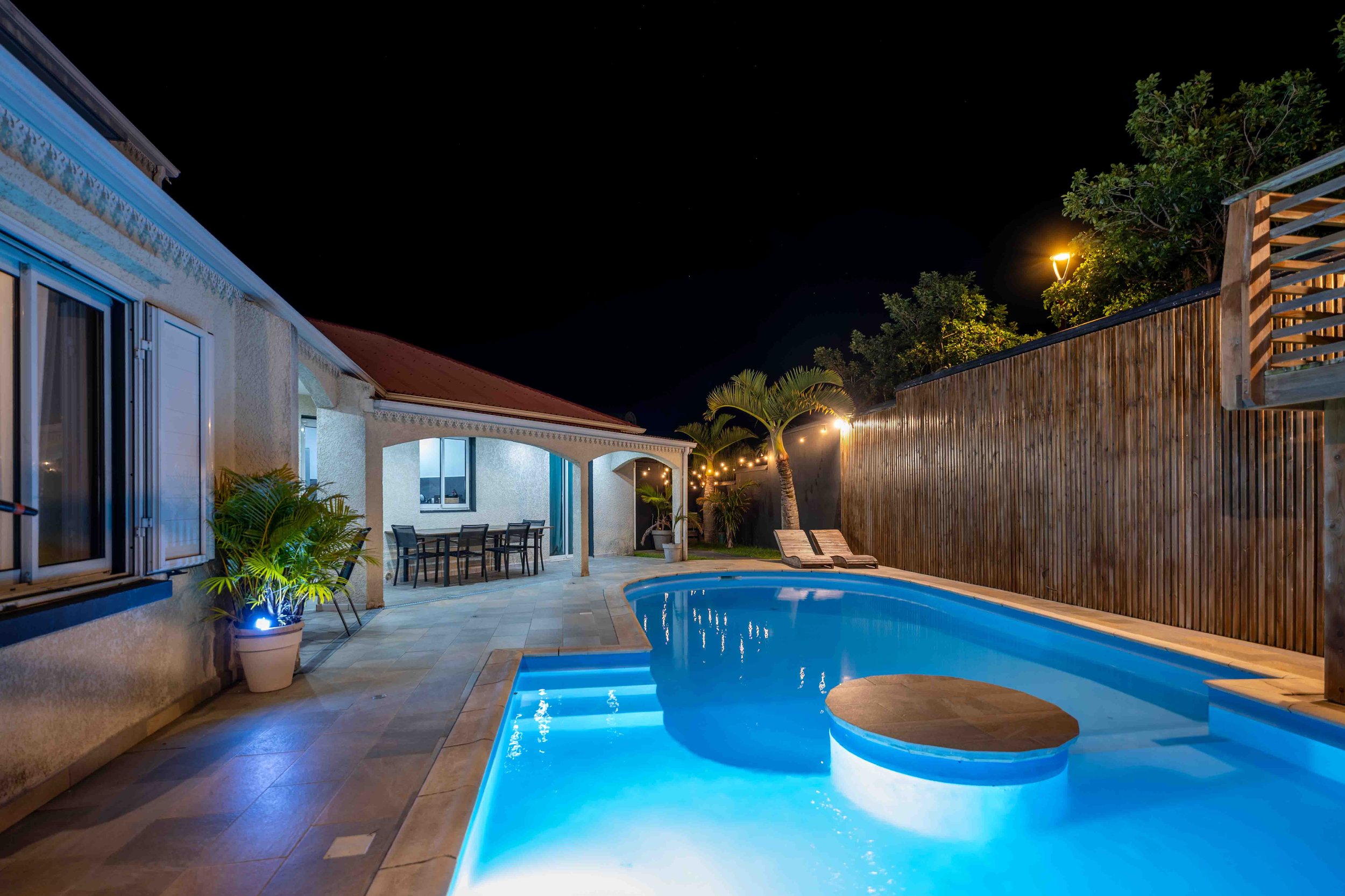 Piscine éclairée la nuit dans une cour arrière, entourée d'une clôture en bois, de palmiers, et d'une maison avec une terrasse, avec des chaises longues et une table de repas. - Photographe immobilier à La Réunion