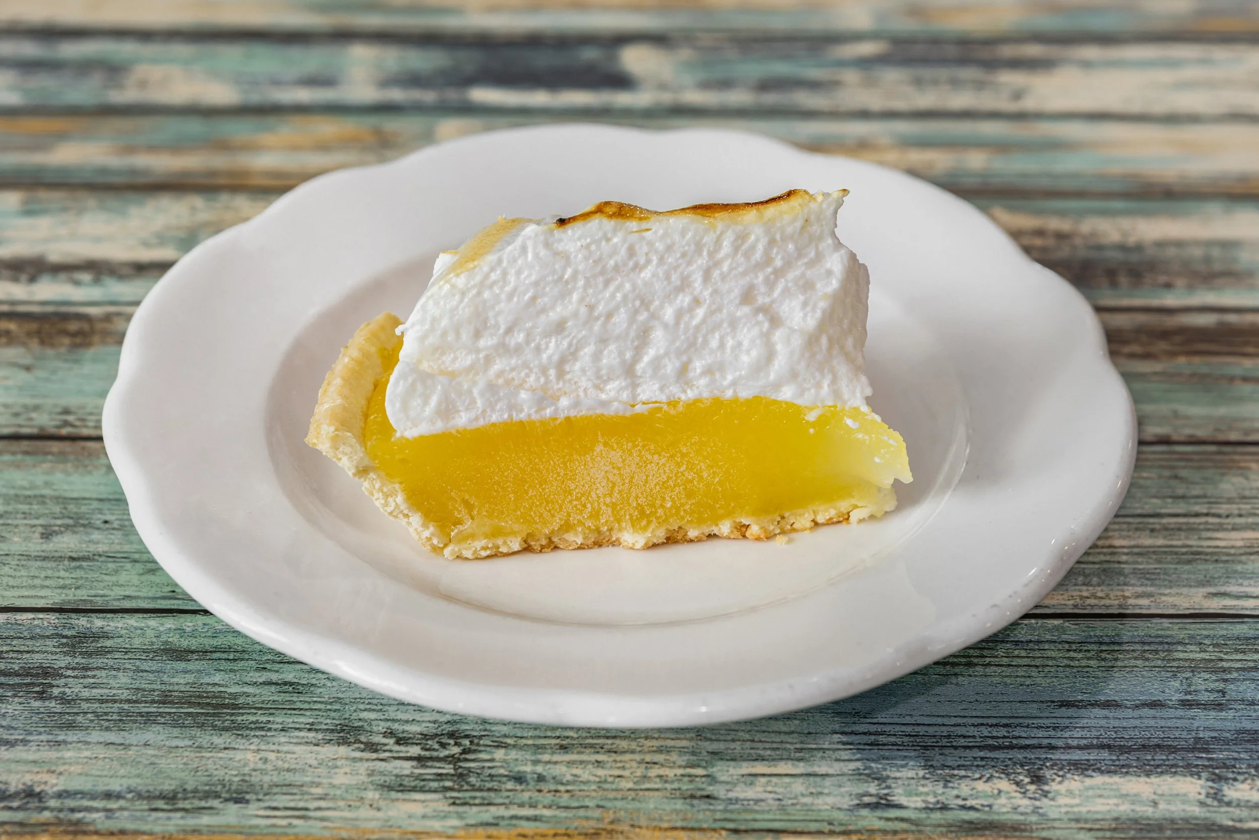 Slice of lemon meringue pie on a white plate, on a rustic wooden table.