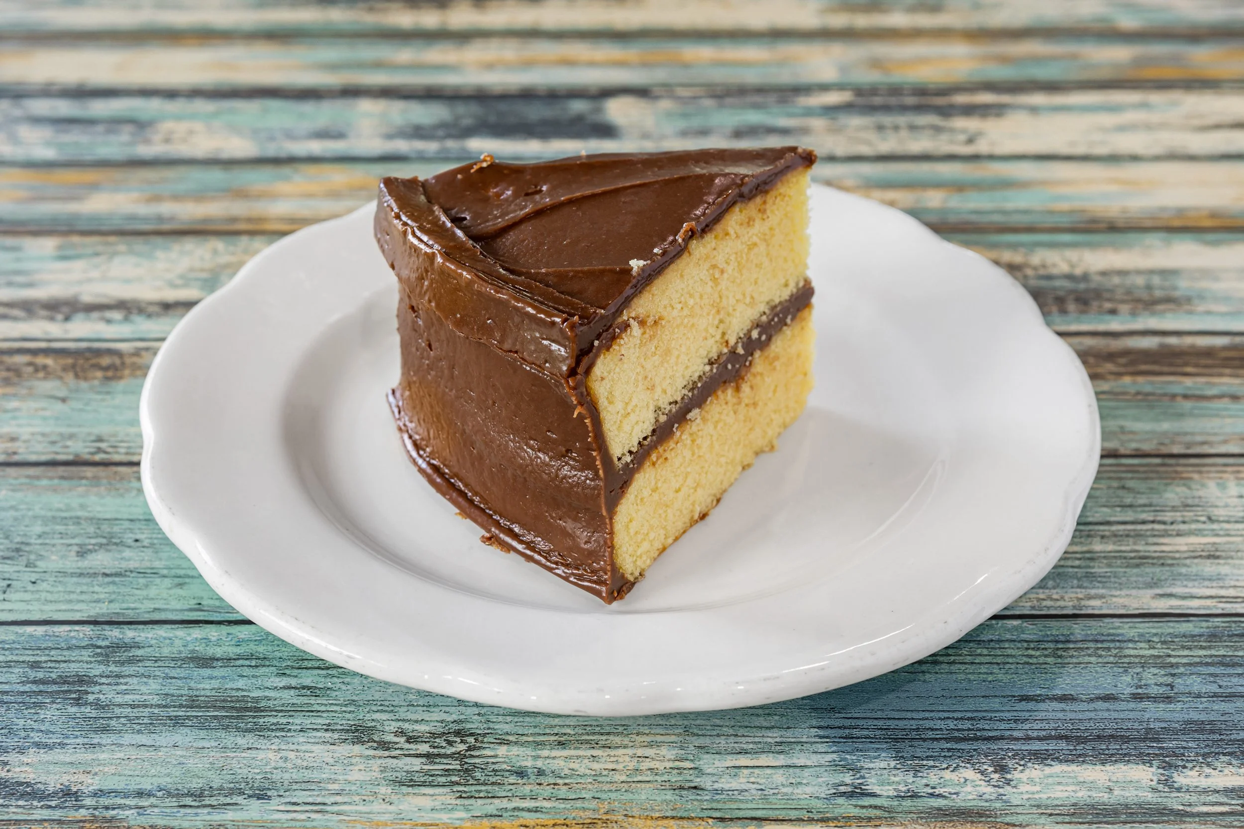 A slice of yellow cake with thick chocolate frosting on a white plate.