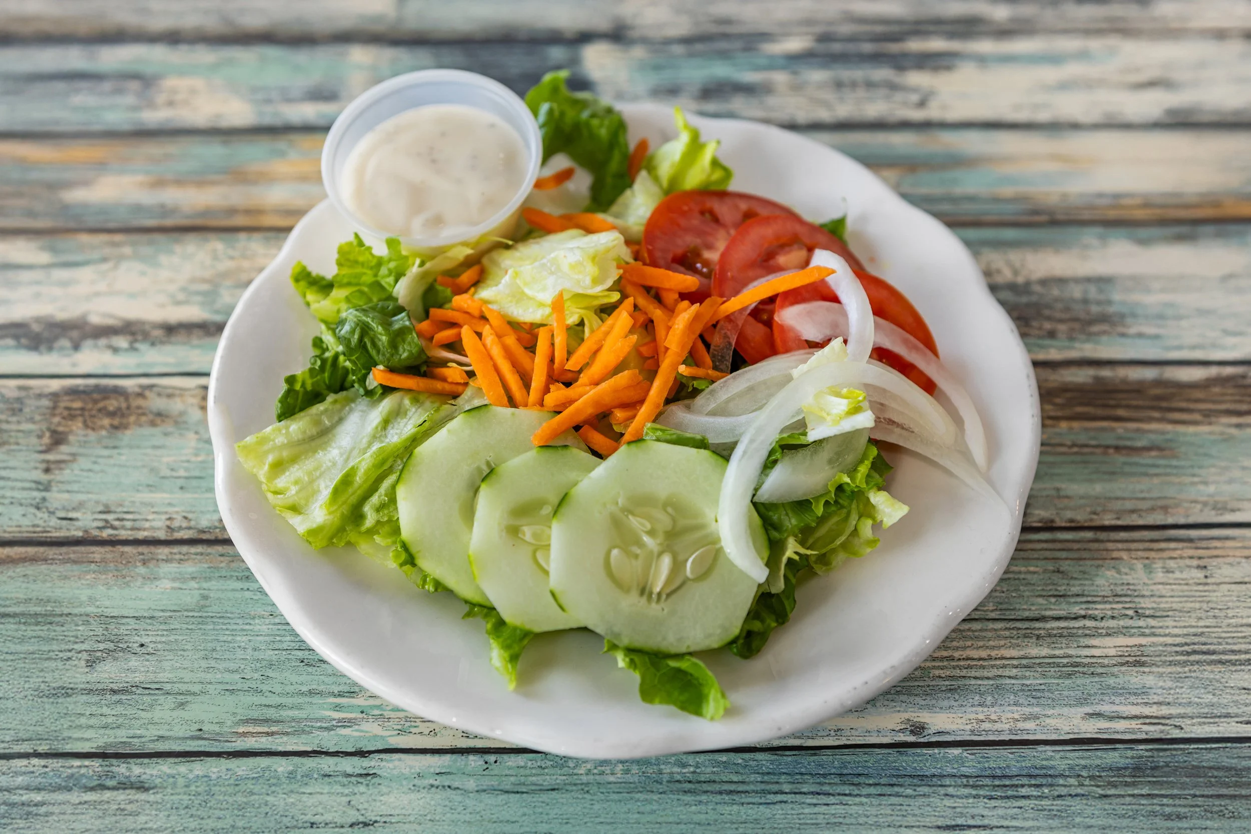 Fresh vegetable salad with cucumbers, tomatoes, onions, shredded carrots, lettuce, and a side of creamy dressing on a white plate, placed on a rustic wooden table.