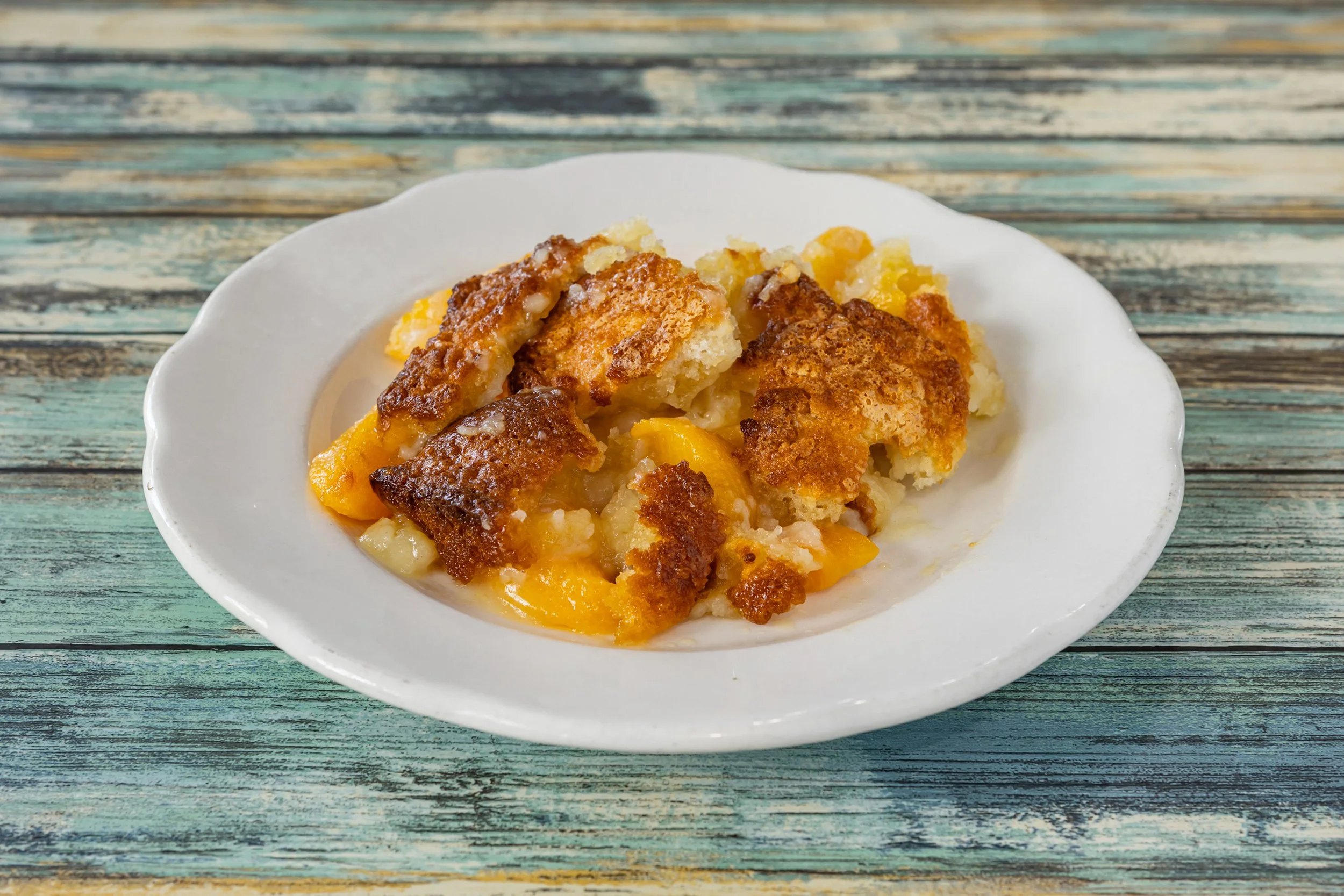 Close-up of a serving of peach cobbler on a white plate, placed on a distressed blue wooden surface.