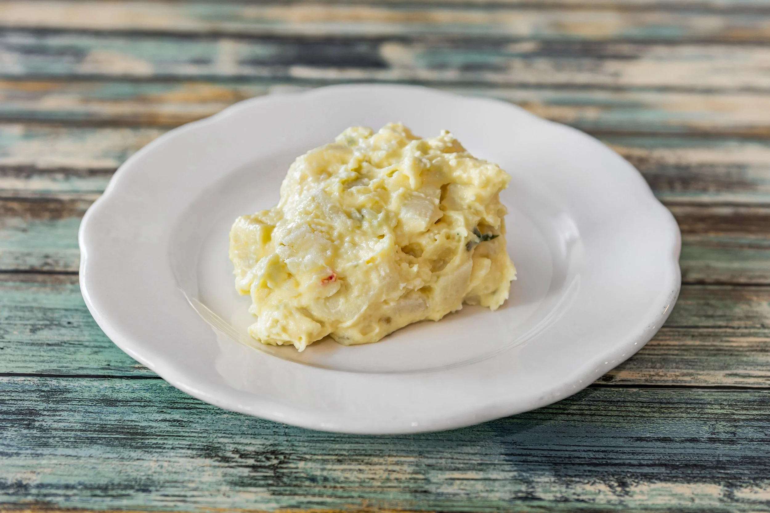 A serving of potato salad on a white decorative plate, placed on a rustic wooden surface.