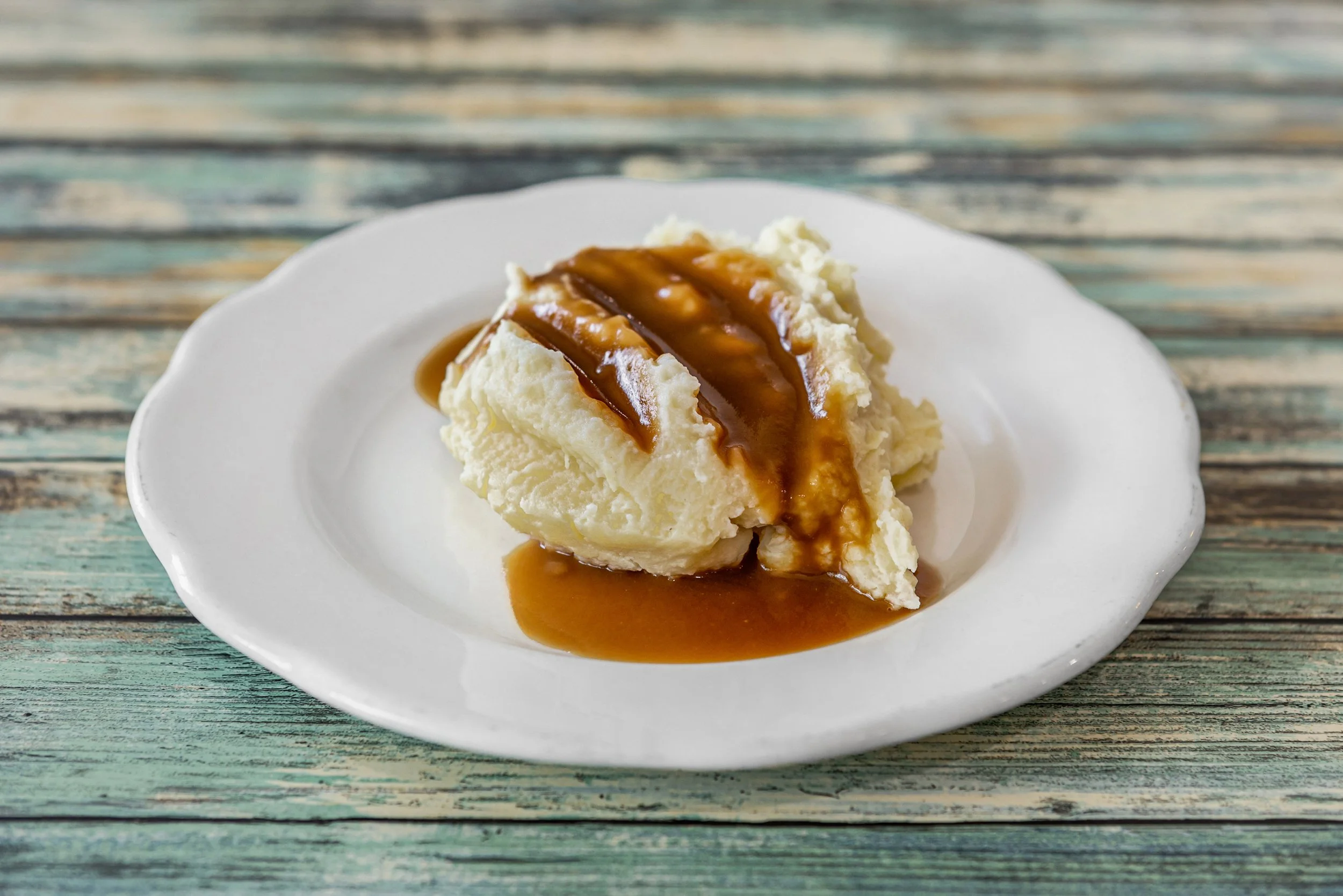 A slice of creamy mashed potatoes topped with rich brown gravy on a white plate with a scalloped edge, placed on a rustic wooden table.