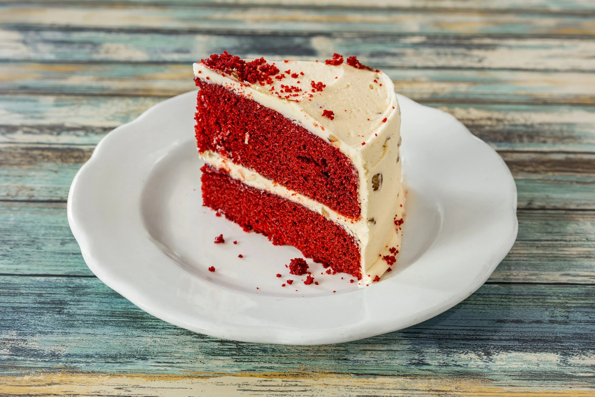 A slice of red velvet cake with cream cheese frosting on a white plate, on a rustic wooden table.