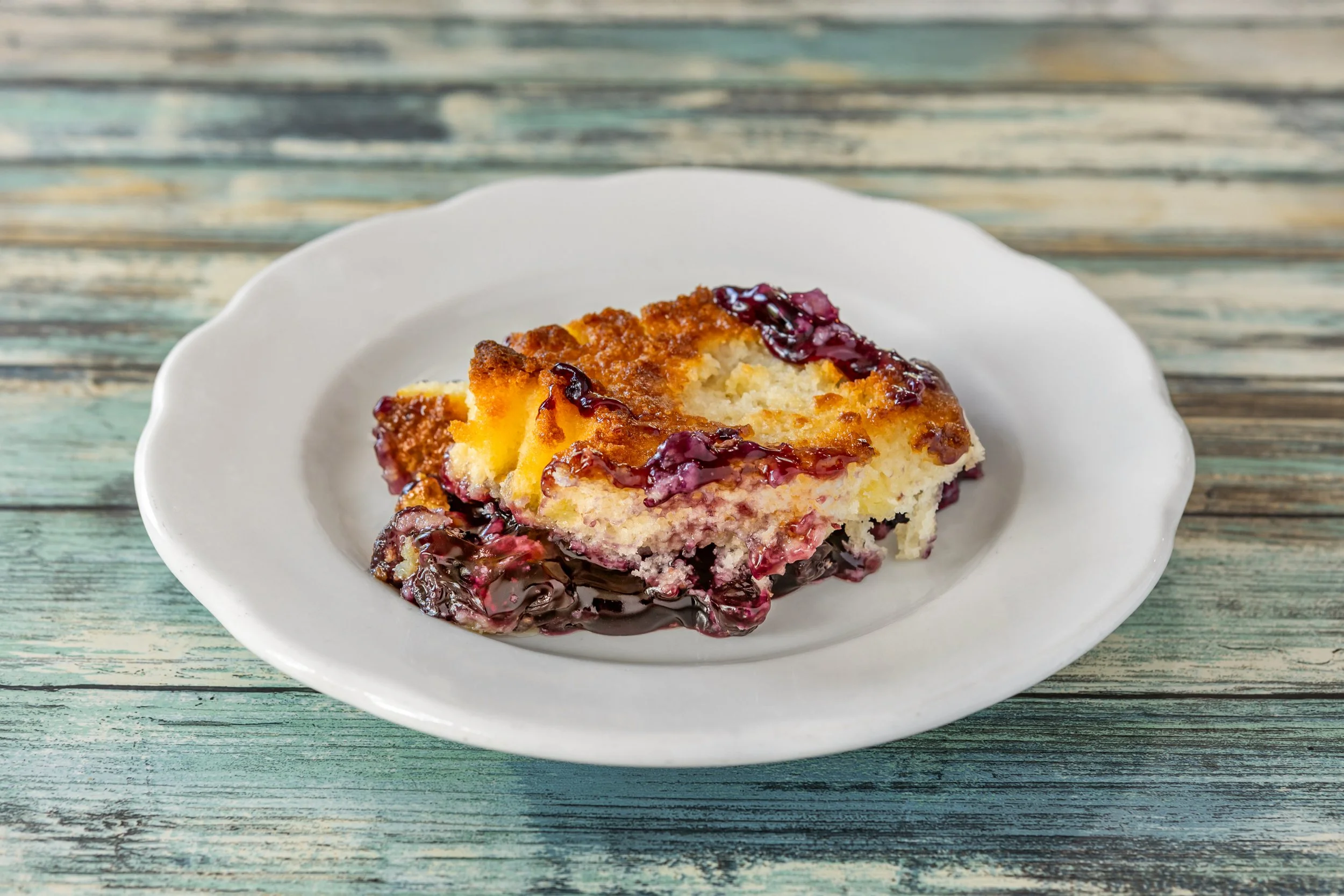 A slice of blueberry cheesecake with blueberry topping on a white scalloped plate, placed on a distressed wood table.