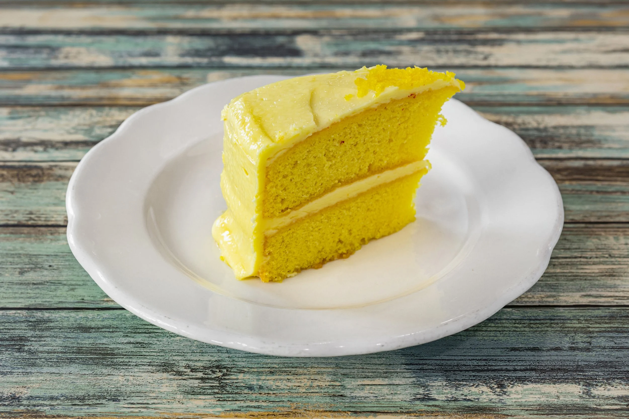 A slice of yellow layered cake with pale yellow frosting on a white plate, on a rustic wooden surface.
